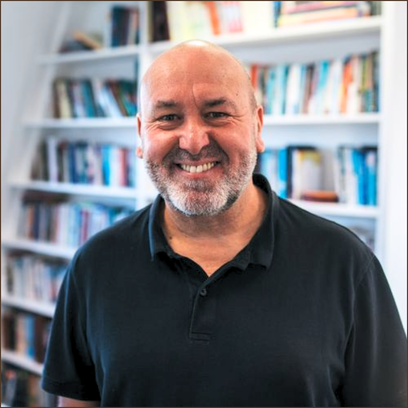 Smiling middle-aged man with a beard wearing a black polo shirt in front of a bookshelf filled with books.