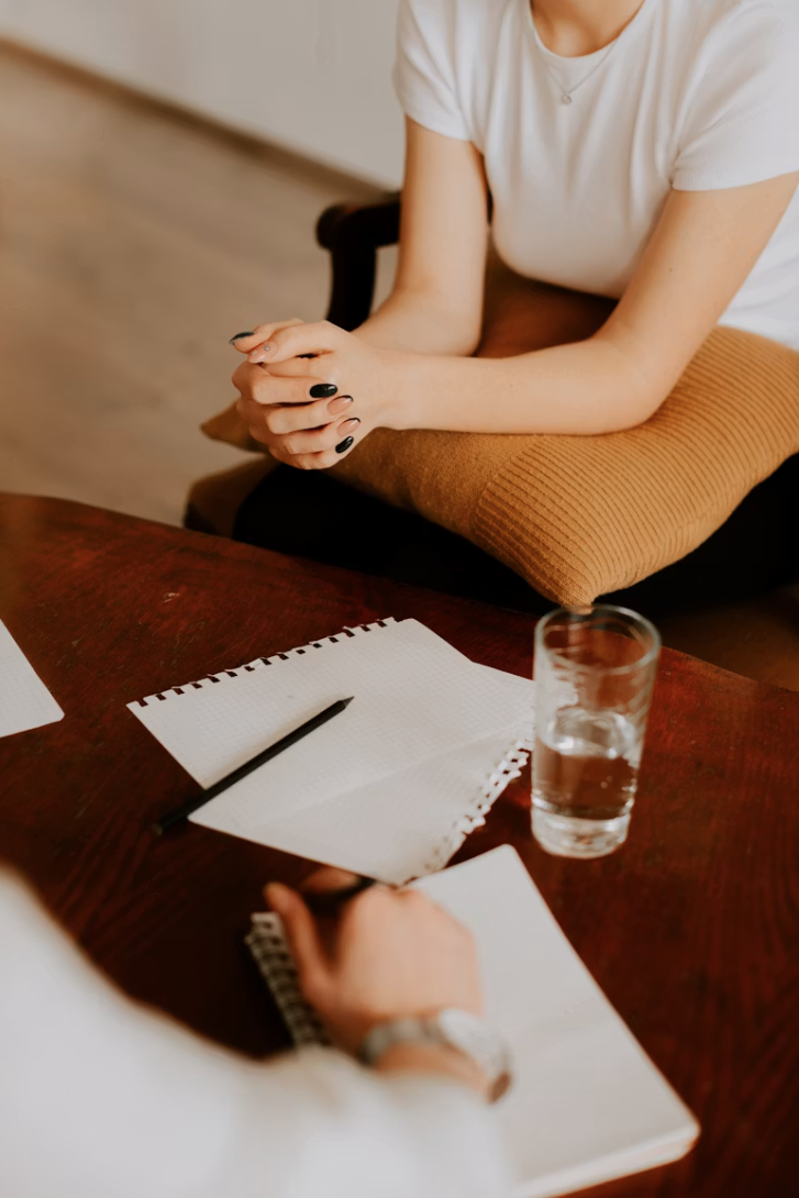 Person sitting at a desk with hands clasped, in front of blank notebooks, a pen, and a glass of water.