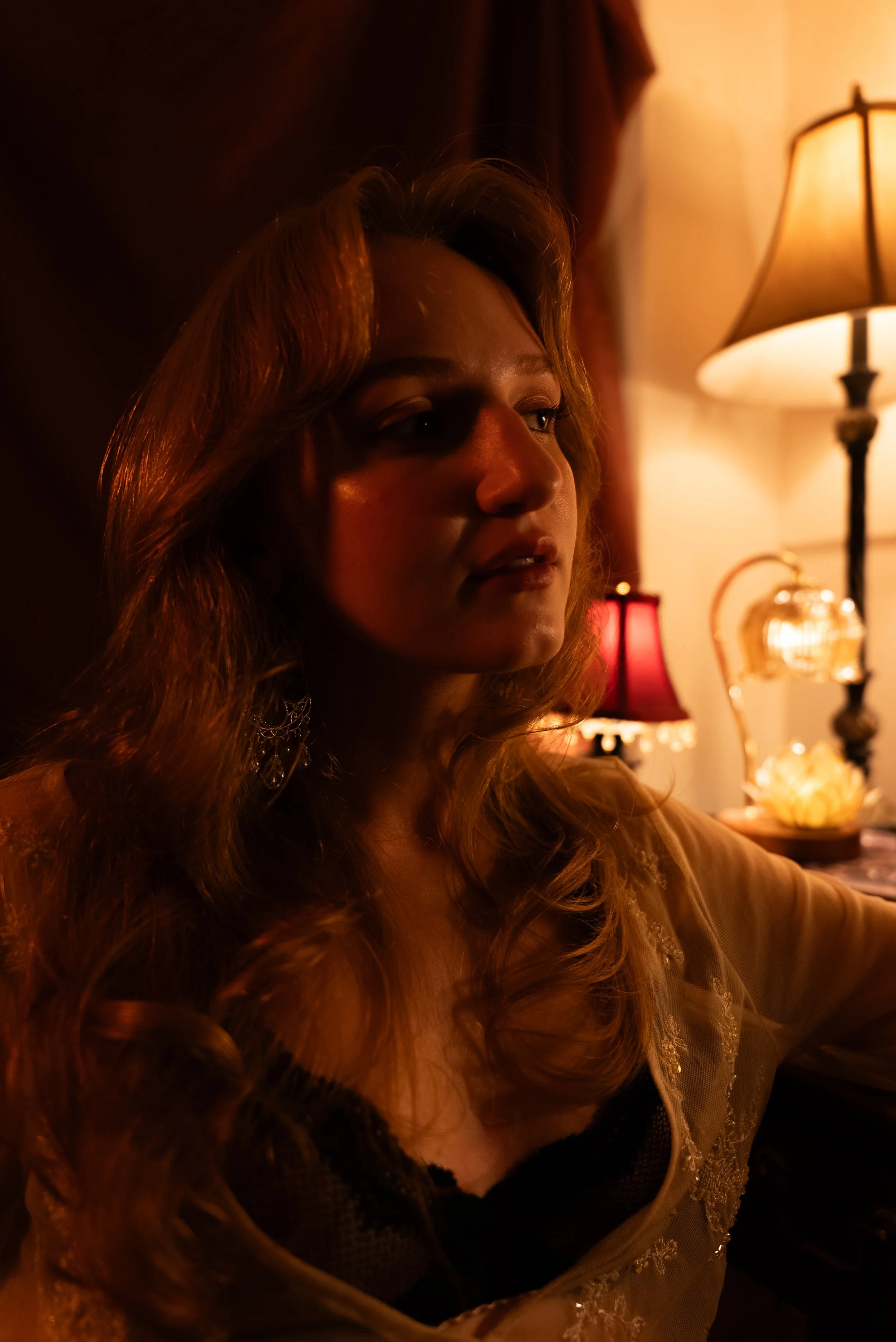 Close portrait of woman in vintage lace with wavy hair lit by warm vintage lamp glow, expression pensive and direct, red lamp visible in background