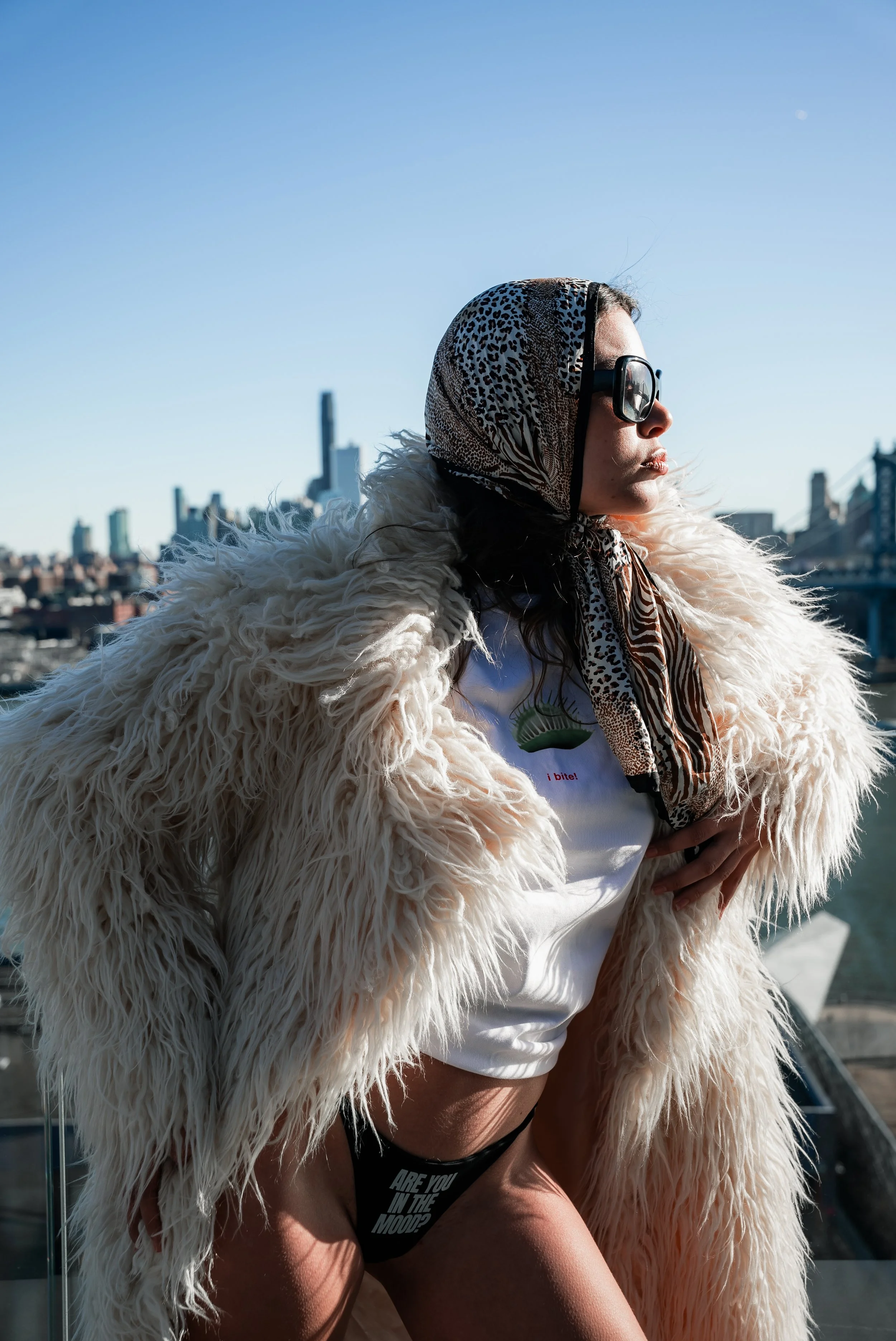 Woman in oversized shearling coat and animal print headscarf gazes off-frame posing on NYC rooftop with city skyline behind her