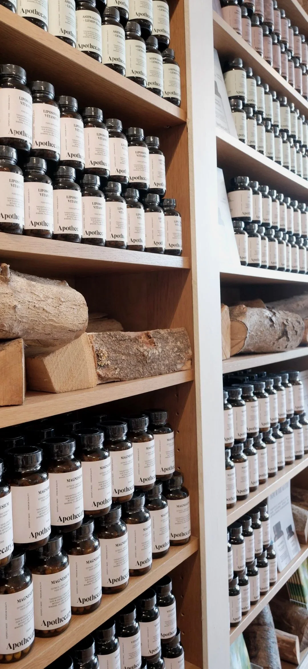 Shelves stocked with brown bottles labeled with ingredients like Ashwagandha Lemon and Magnesium, wood logs on lower shelves, in an apothecary or herbal store.