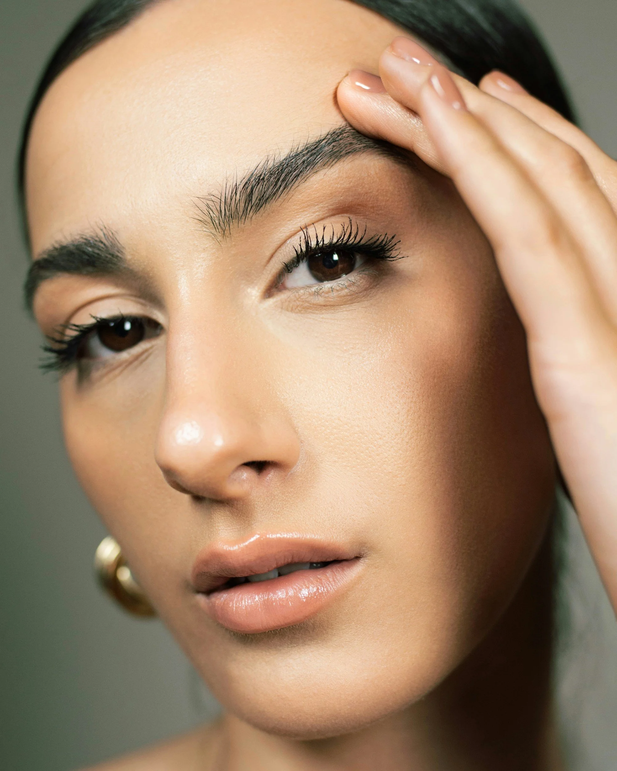 Close-up of a woman's face, showing her eye, eyebrow, and part of her forehead. She has dark hair, well-groomed eyebrows, and is touching her forehead with her hand. Her makeup appears natural with glossy lips.