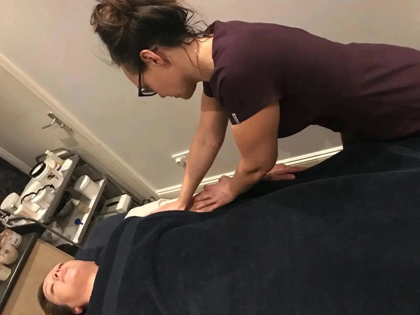 A woman wearing glasses and a maroon shirt performing a massage on a person lying face down on a bed in a room with beige walls and medical supplies visible on a shelf.