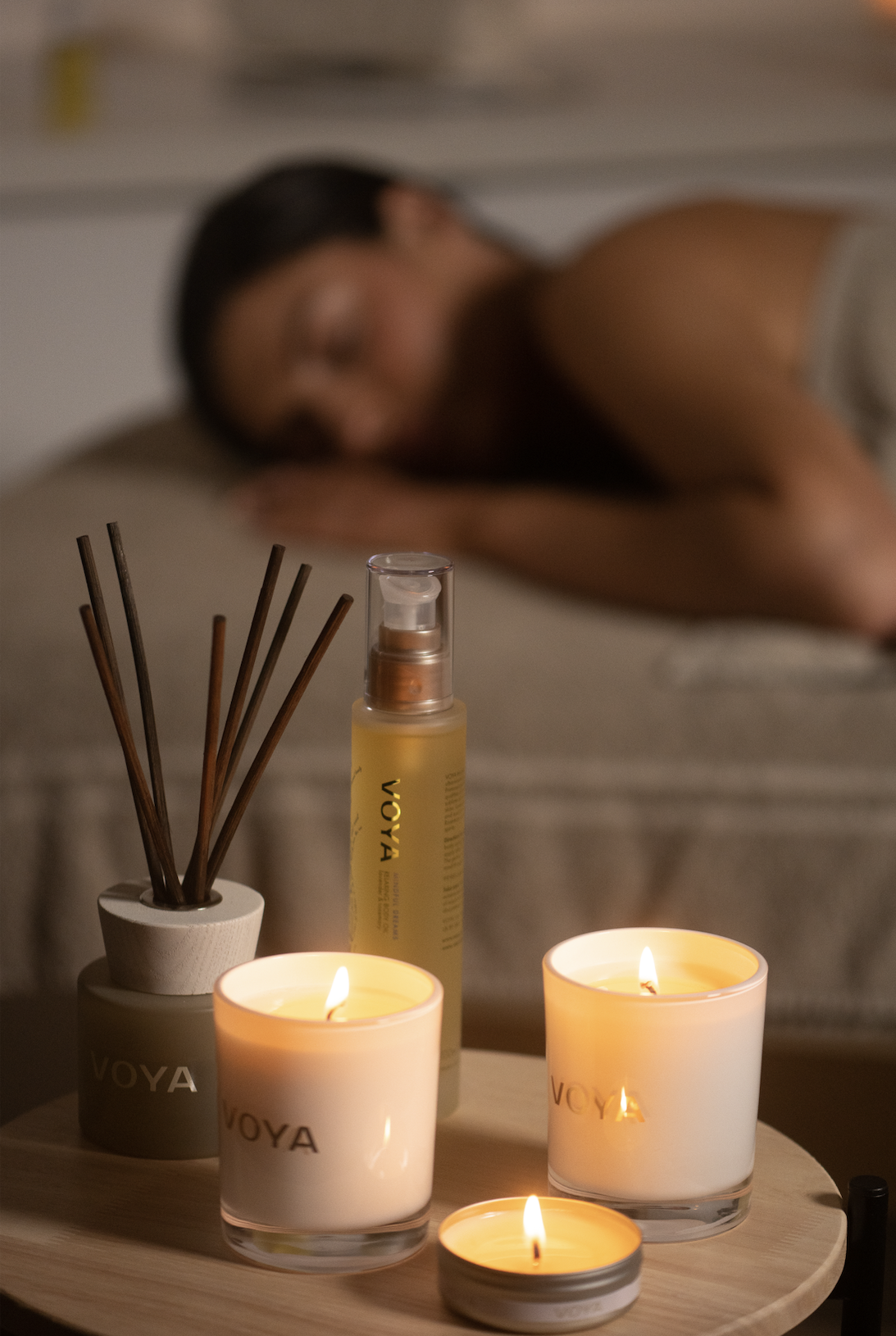 A woman lies on her side with her eyes closed in a bedroom, with a wooden table in the foreground holding lit candles, a spray bottle, and reed diffusers.