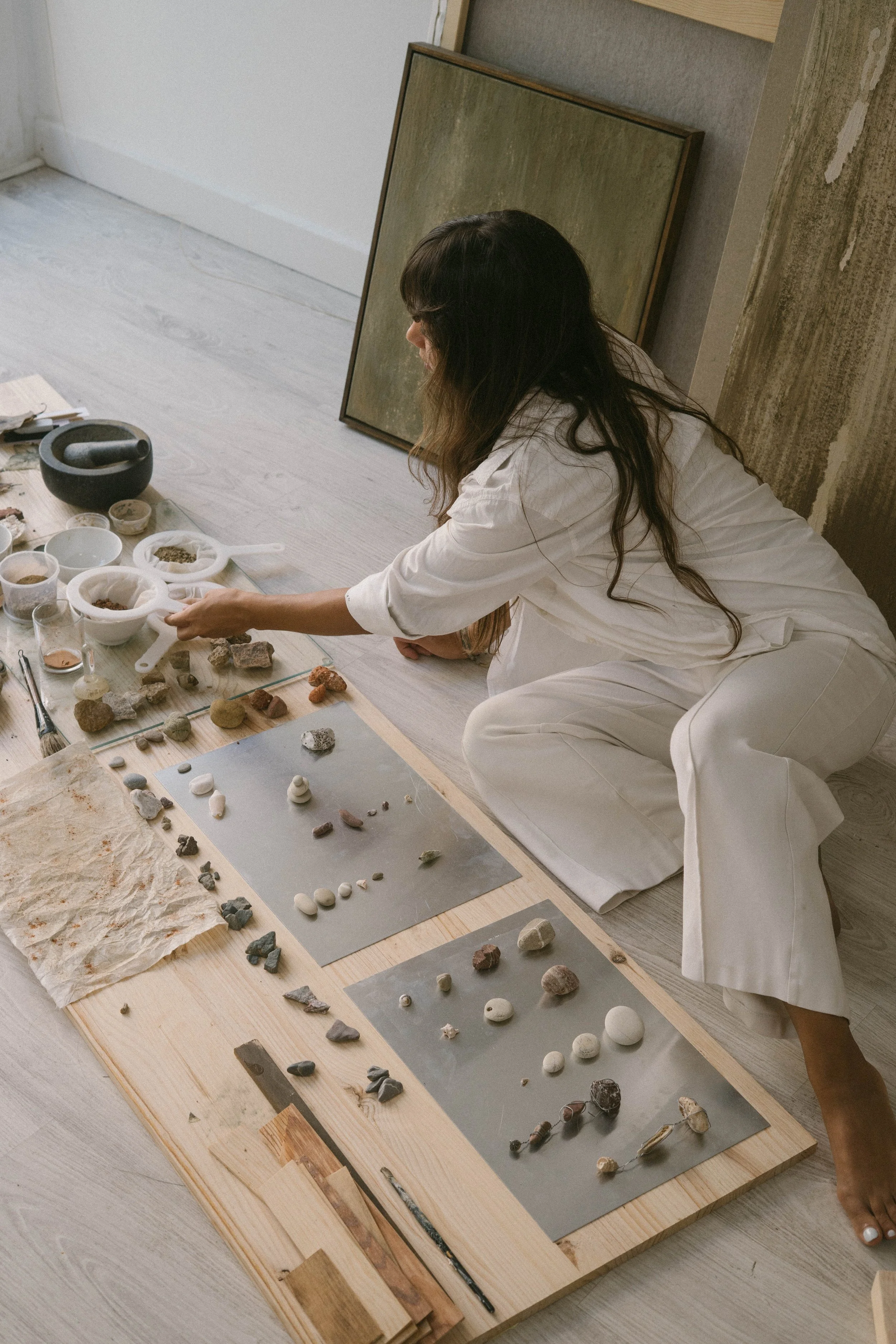 A woman in white clothing sitting on the floor, examining a collection of rocks and minerals organized on slabs and in bowls, with framed artworks leaning against the wall beside her.