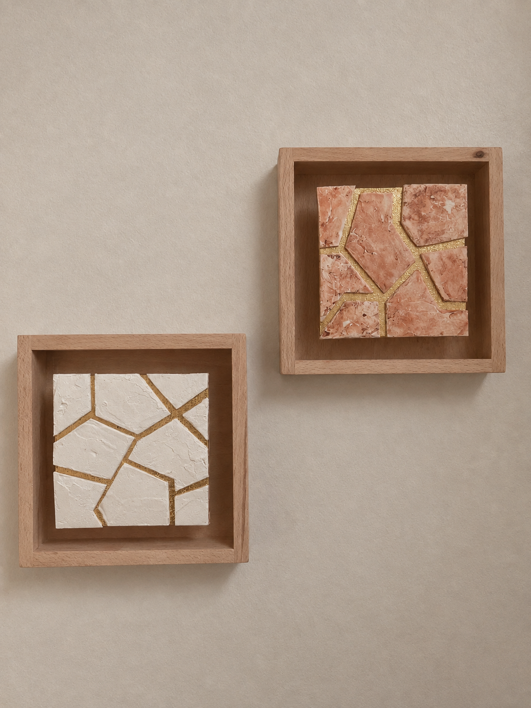 Two wooden shadow boxes containing mosaic tiles, one white and one pinkish-orange, arranged in geometric broken patterns, on a beige surface.