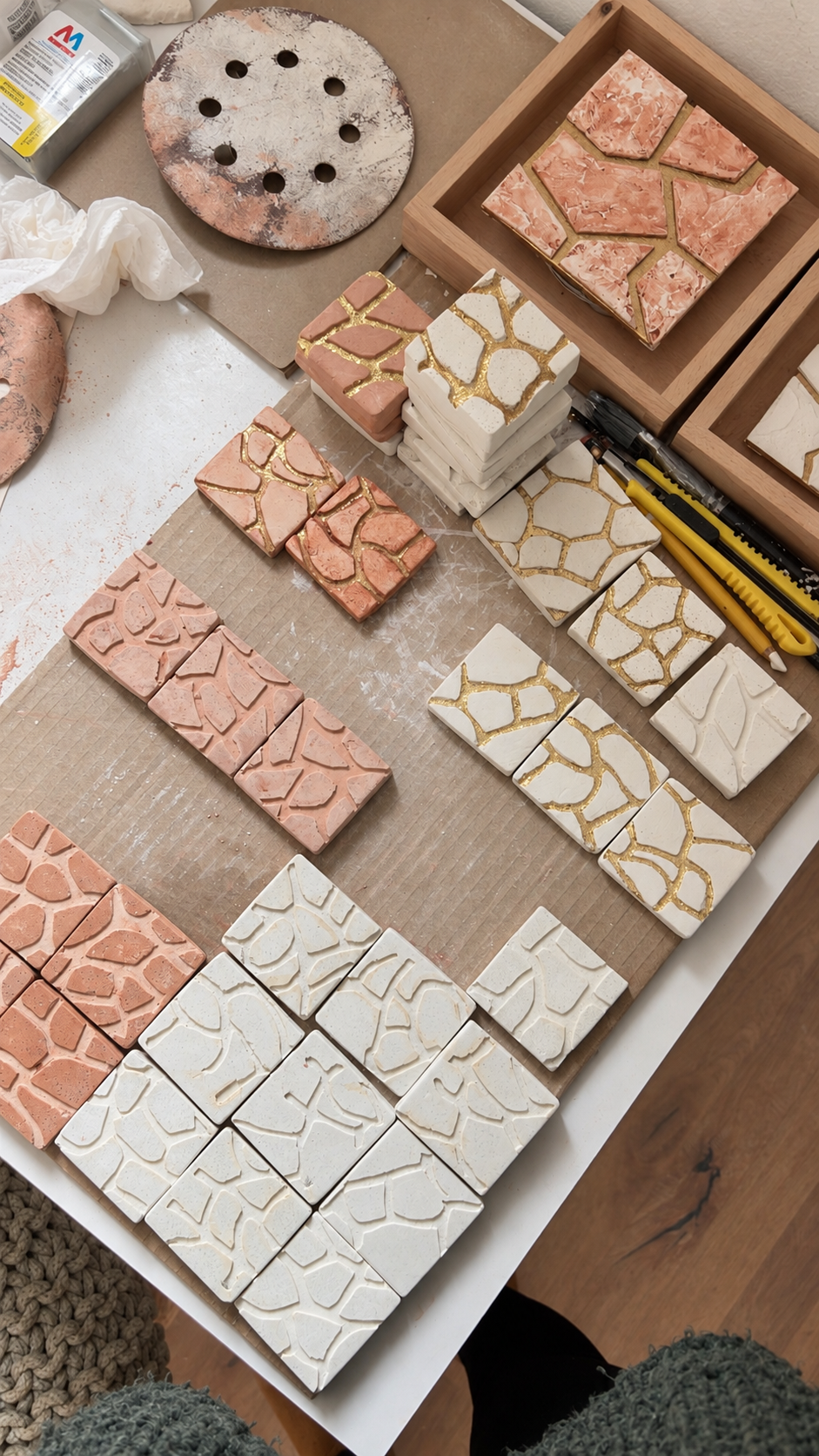 Ceramic tiles with a mosaic pattern, some with pink, white, and gold details, arranged on a work surface with tools and boxes nearby.