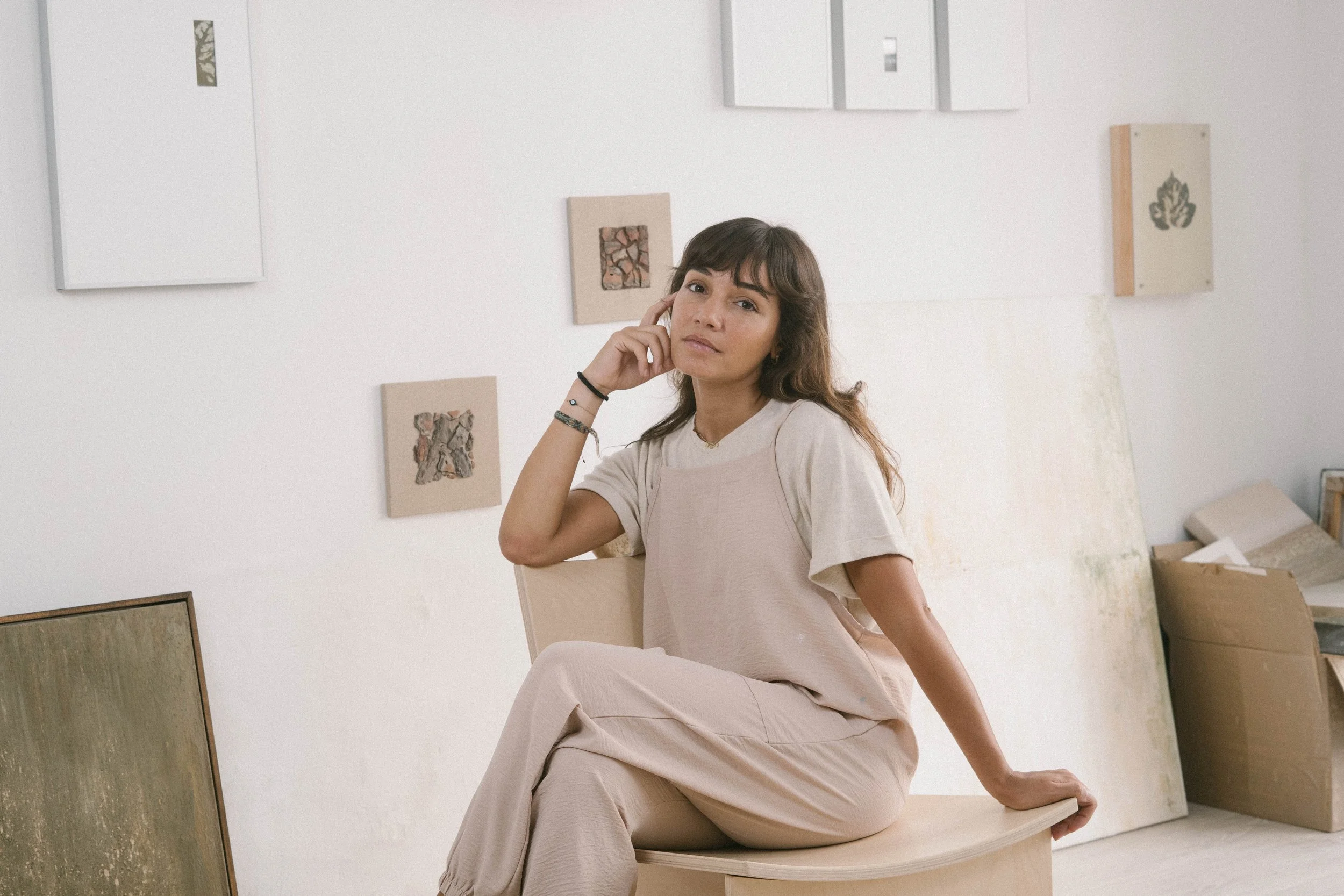 A woman with brown hair sitting on a wooden stool in an art studio, surrounded by abstract artwork and paintings leaning against the wall.