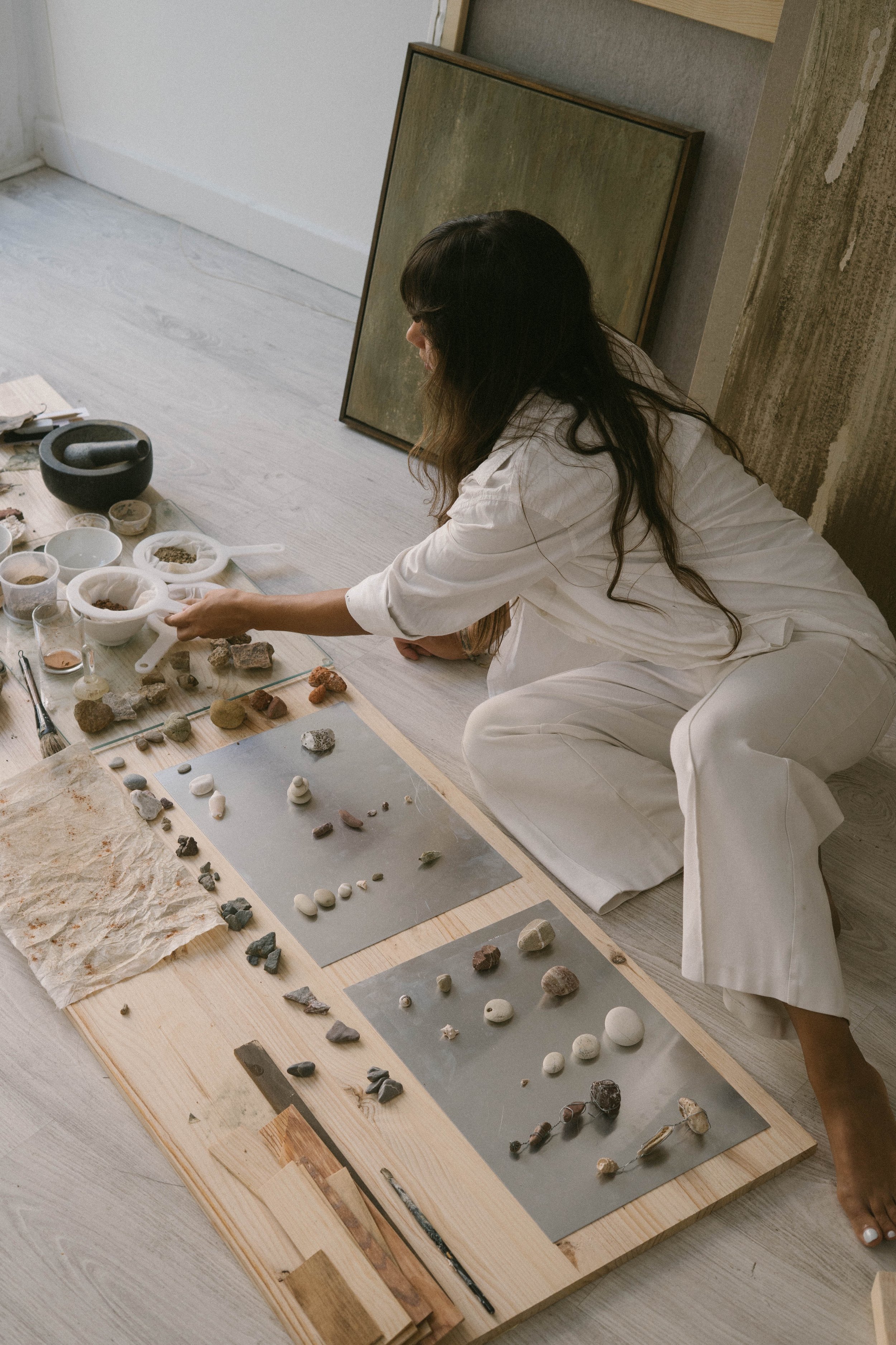 A woman with long dark hair, dressed in white, is lying on the floor examining various rocks and minerals arranged on wooden and gray mats.