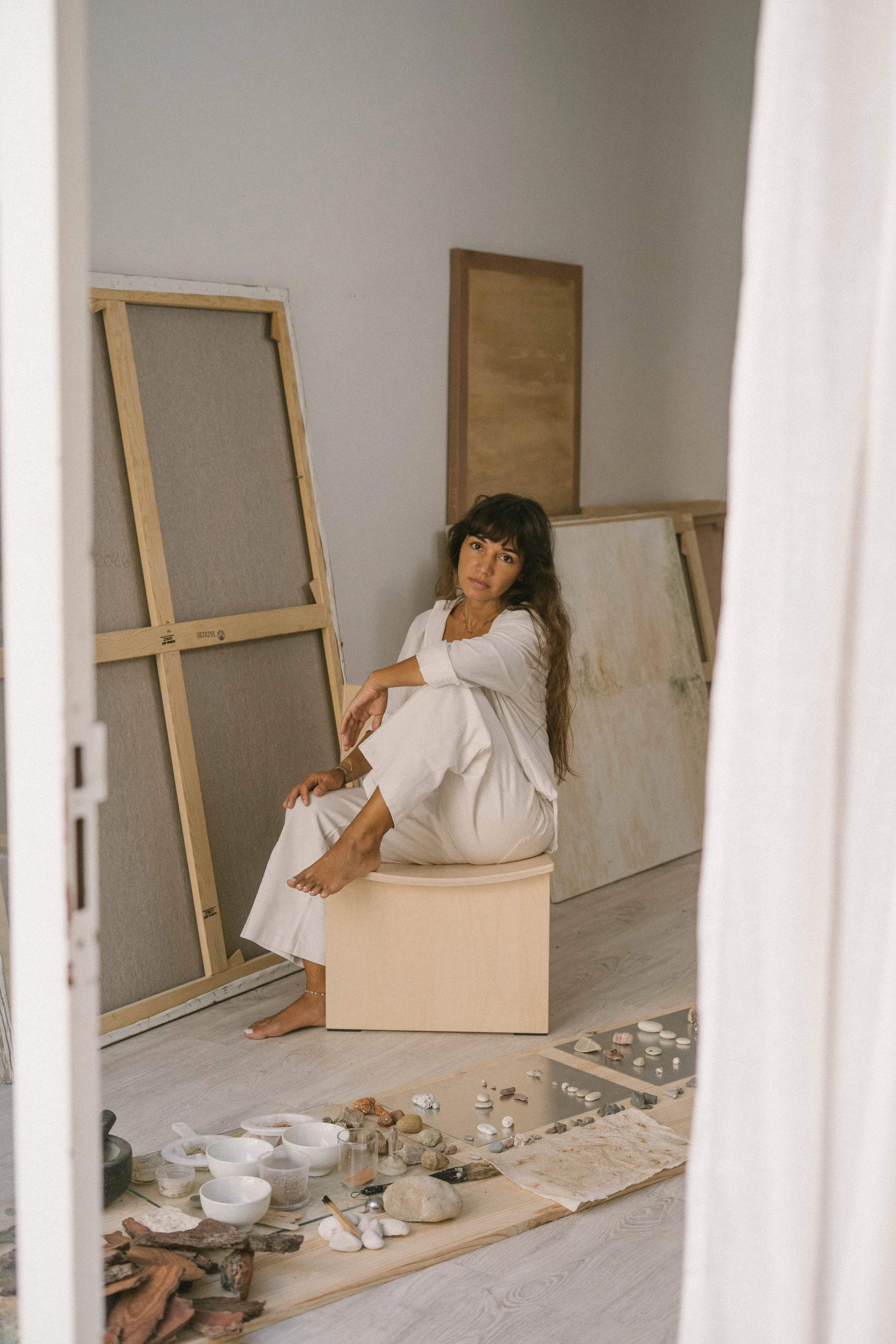 A woman sits barefoot on a round stool in an art studio, surrounded by canvases, stones, shells, and pottery arranged on a mat and wooden board on the floor, as she looks at the camera with a relaxed expression.