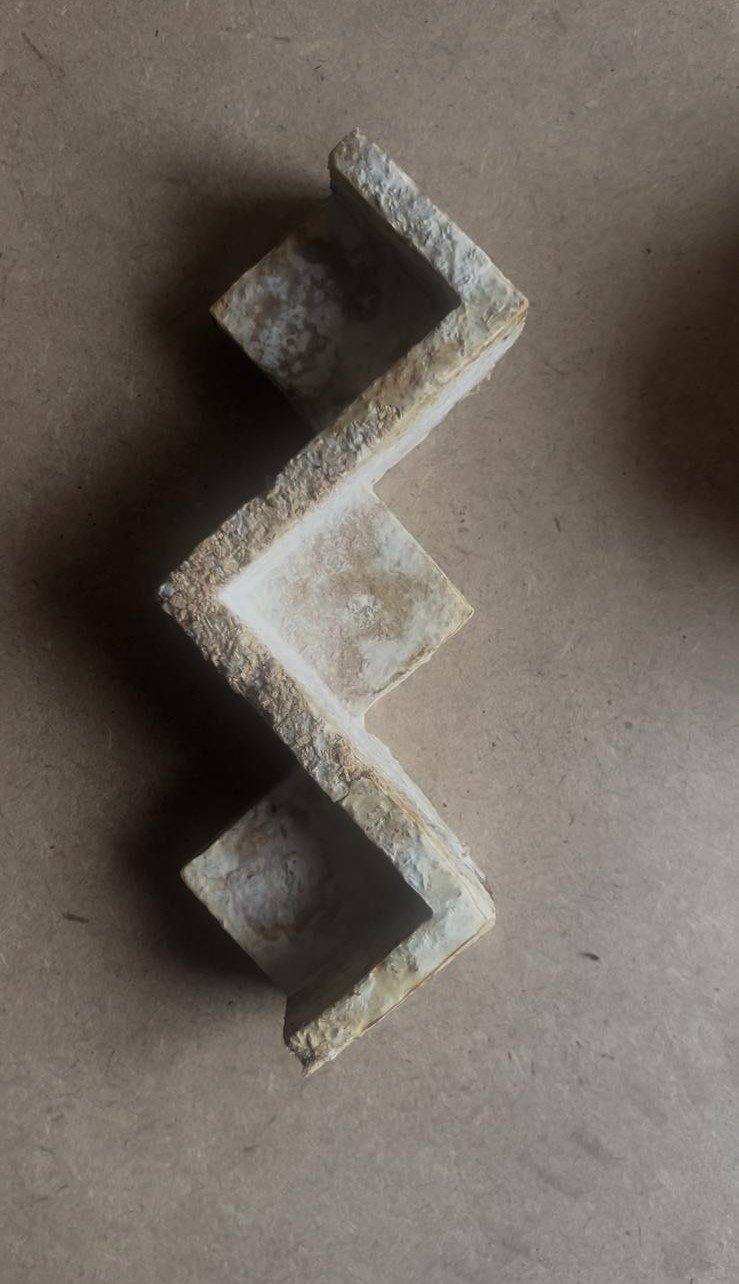 A zigzag-shaped metal or concrete piece with a rough, weathered surface, placed on a plain, brown background.
