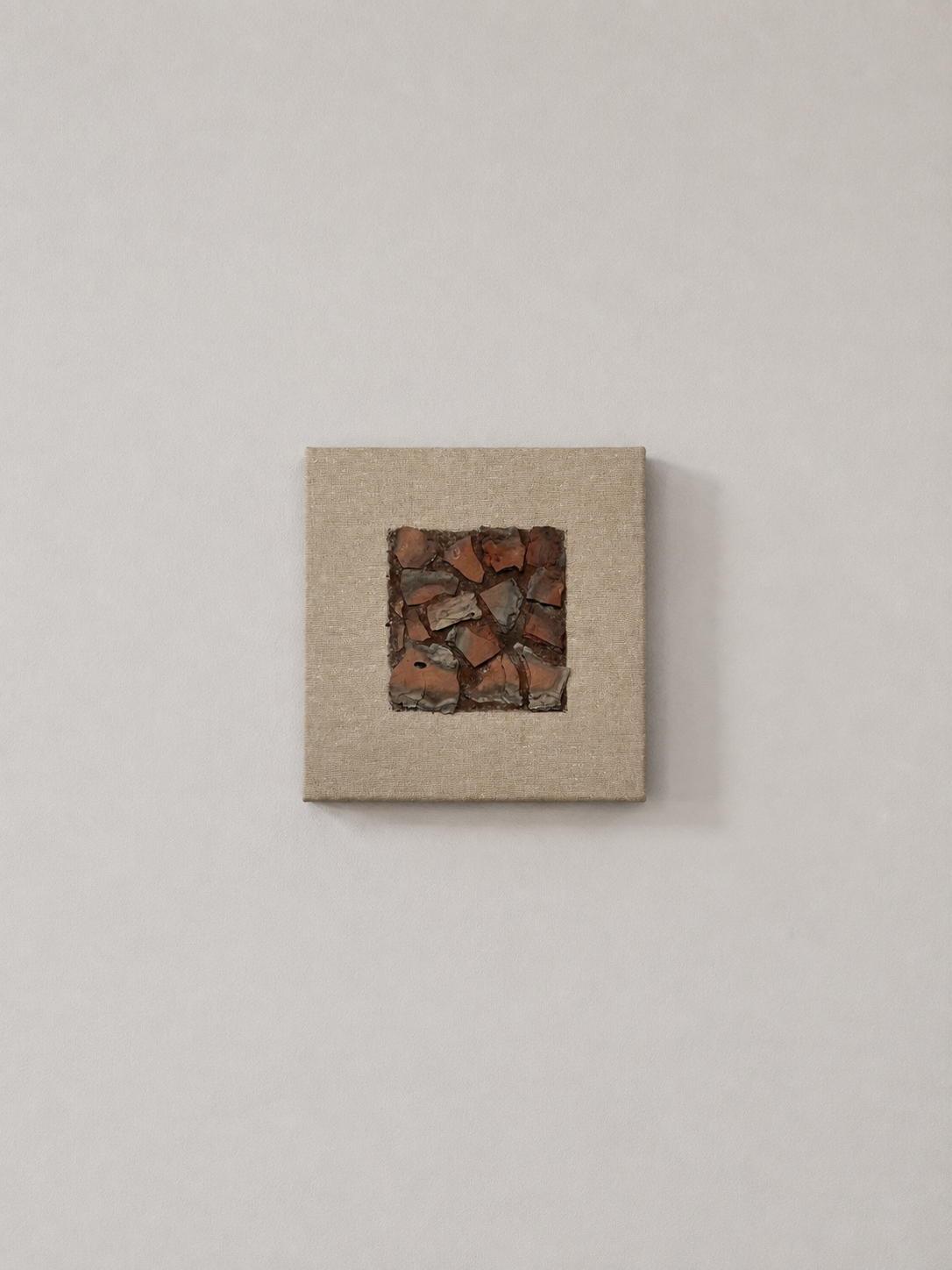 Contemporary art piece with a square of broken terracotta tiles embedded in a beige canvas frame, mounted on a white wall.