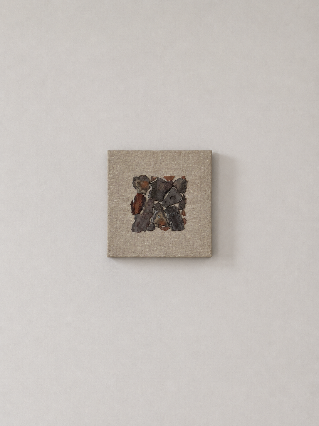 Abstract artwork featuring rocks and iron fragments mounted on a neutral canvas, hung on a plain white wall.