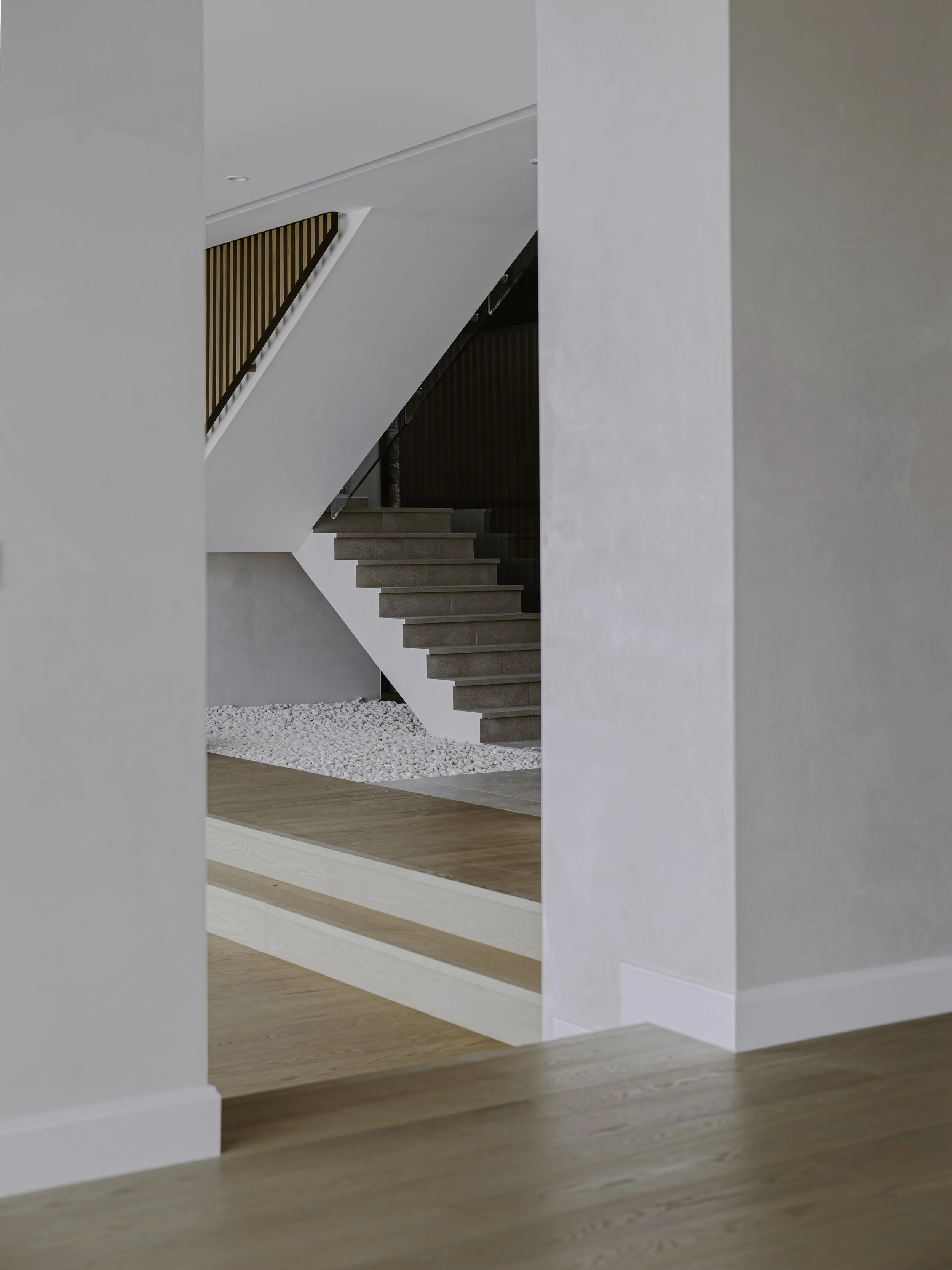 Modern interior staircase with beige steps, wooden railing, glass balustrade, and a white wall with a textured finish. Wood and concrete flooring at the bottom of the stairs.