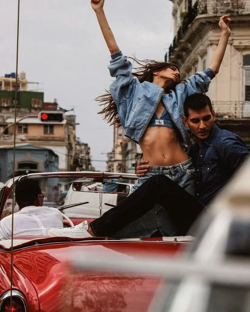 Two young adults, a woman and a man, are sitting in a red convertible car, with the woman standing and raising arms in the air while the man is seated. The woman is wearing a denim jacket and has her midriff exposed. The scene is set on a city street with buildings and traffic in the background.