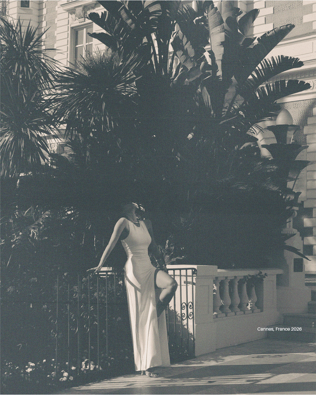 A woman in a long, elegant dress with a high slit, standing outdoors next to a fence, with large tropical plants and a building in the background. Text on the image reads "Cannes, France 2026".