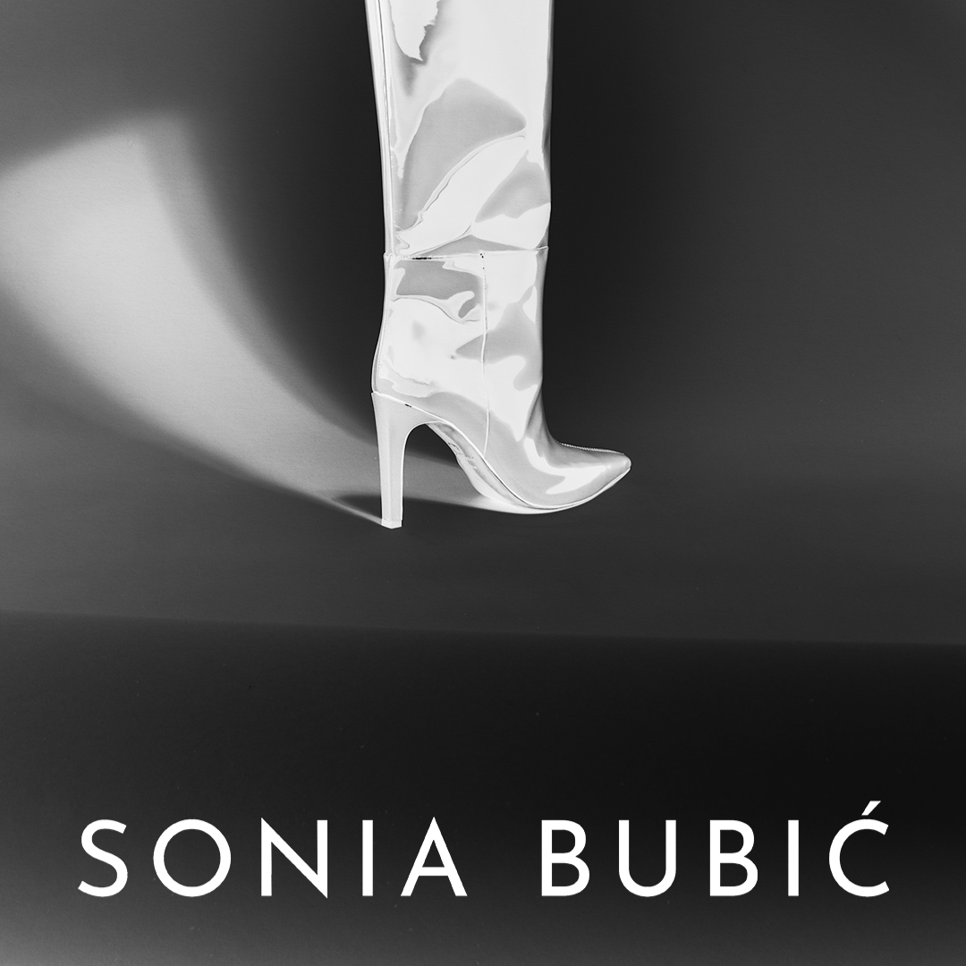 Black and white photo of a shiny, silver high-heeled boot with a pointed toe, set against a dark background with a soft shadow.