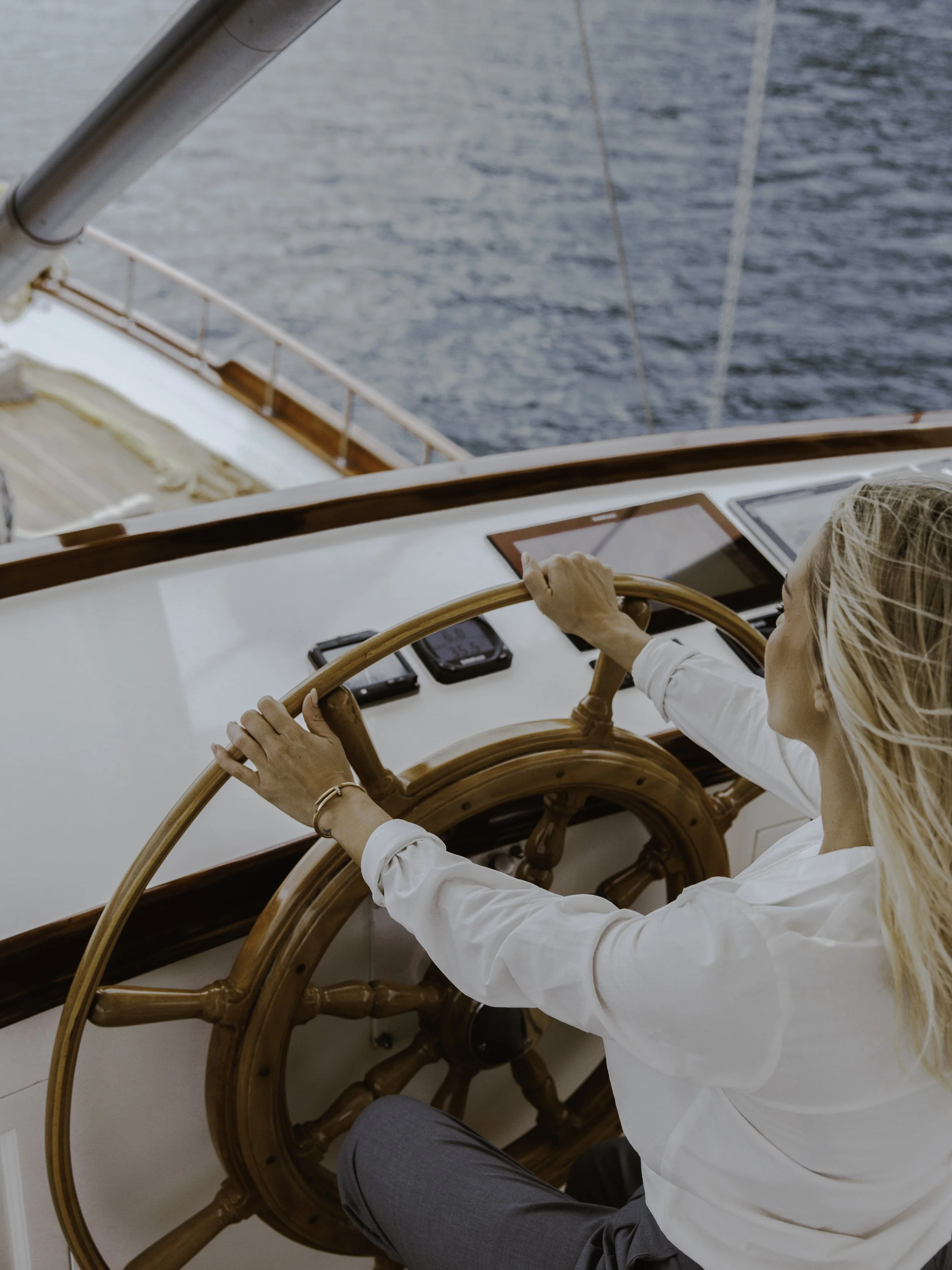 A woman steering a boat at sea, holding the wheel with her right hand and using a navigation device with her left hand, with water visible outside.