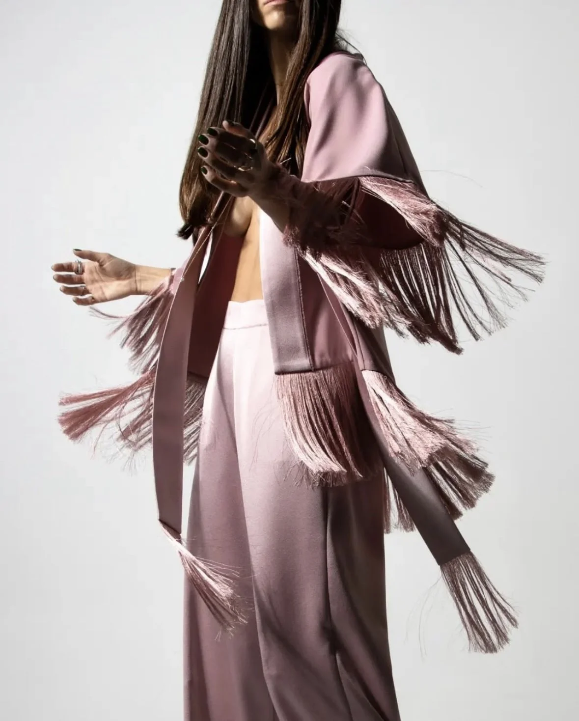 A woman wearing a pink satin outfit with fringed sleeves and wide-leg pants, standing against a plain background.