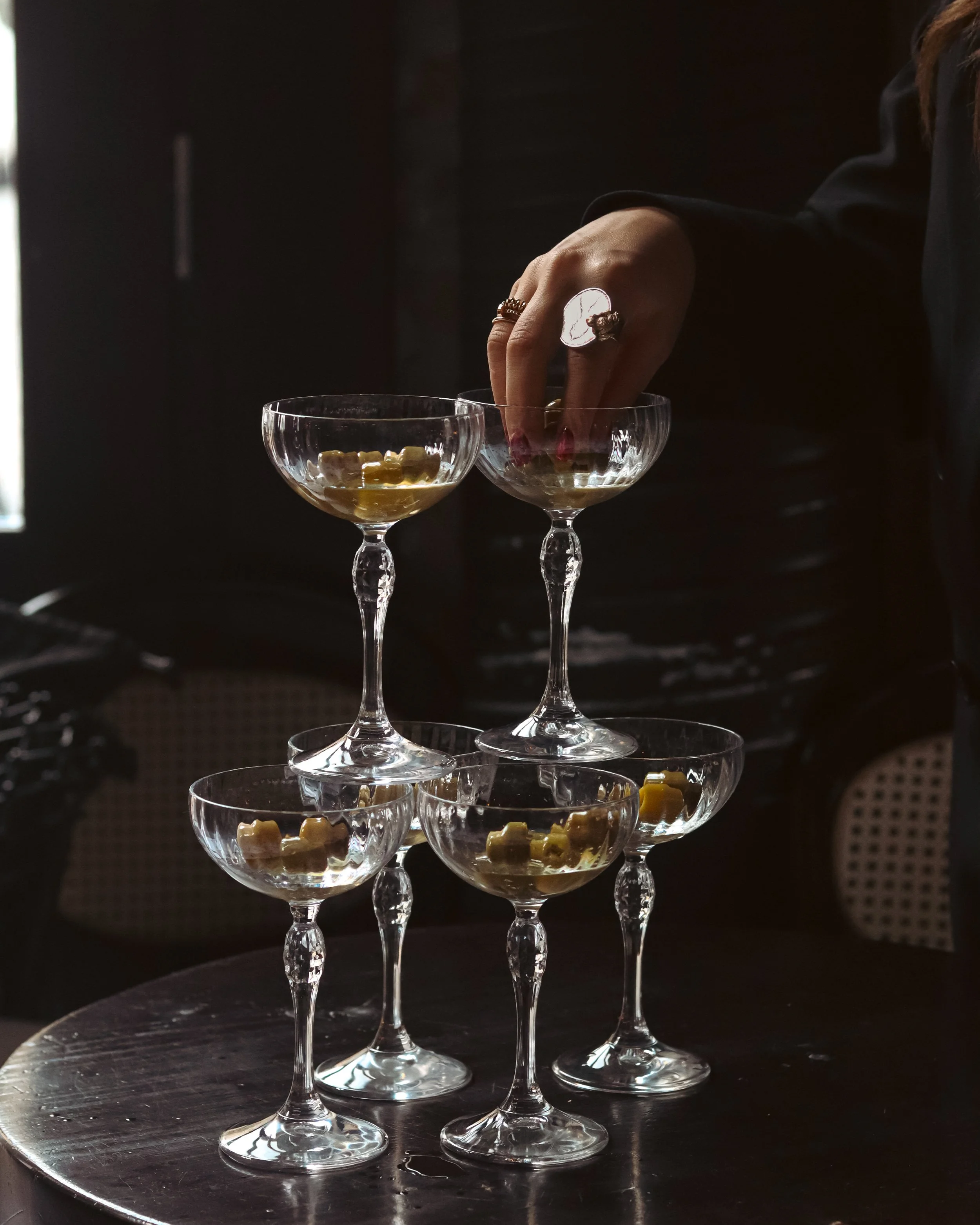 A person's hand with rings encrusted with jewels is reaching into a glass bowl filled with small pieces of cheese, standing among several other empty glass bowls stacked on a dark round surface, with a dark background and reflection on the surface.