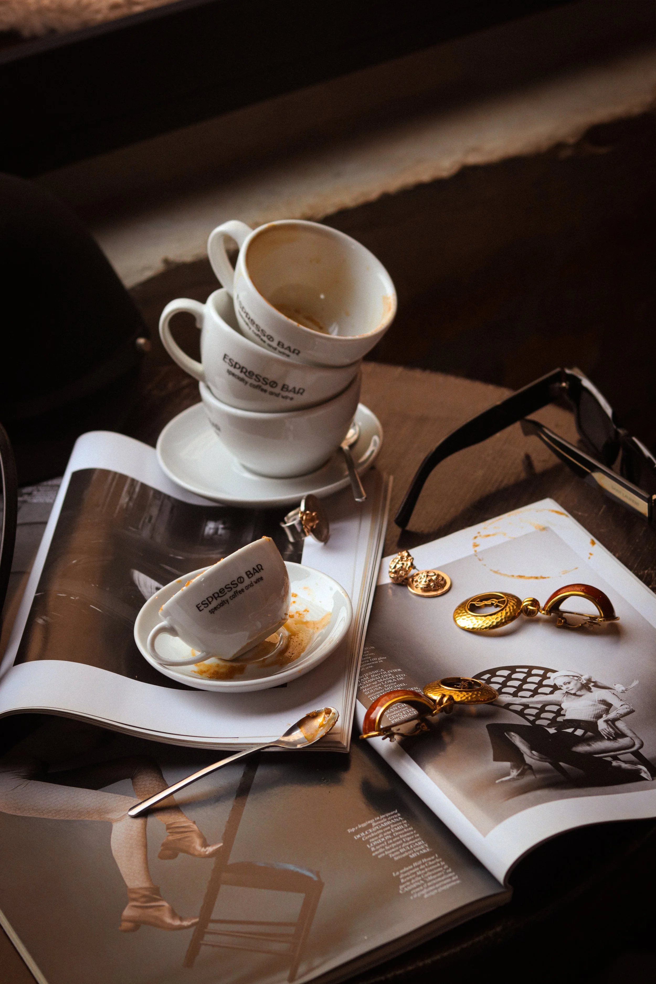 Three white espresso cups with coffee remnants on saucers stacked on top of each other, placed on an open magazine with black and white images and gold jewelry, on a dark wooden surface.