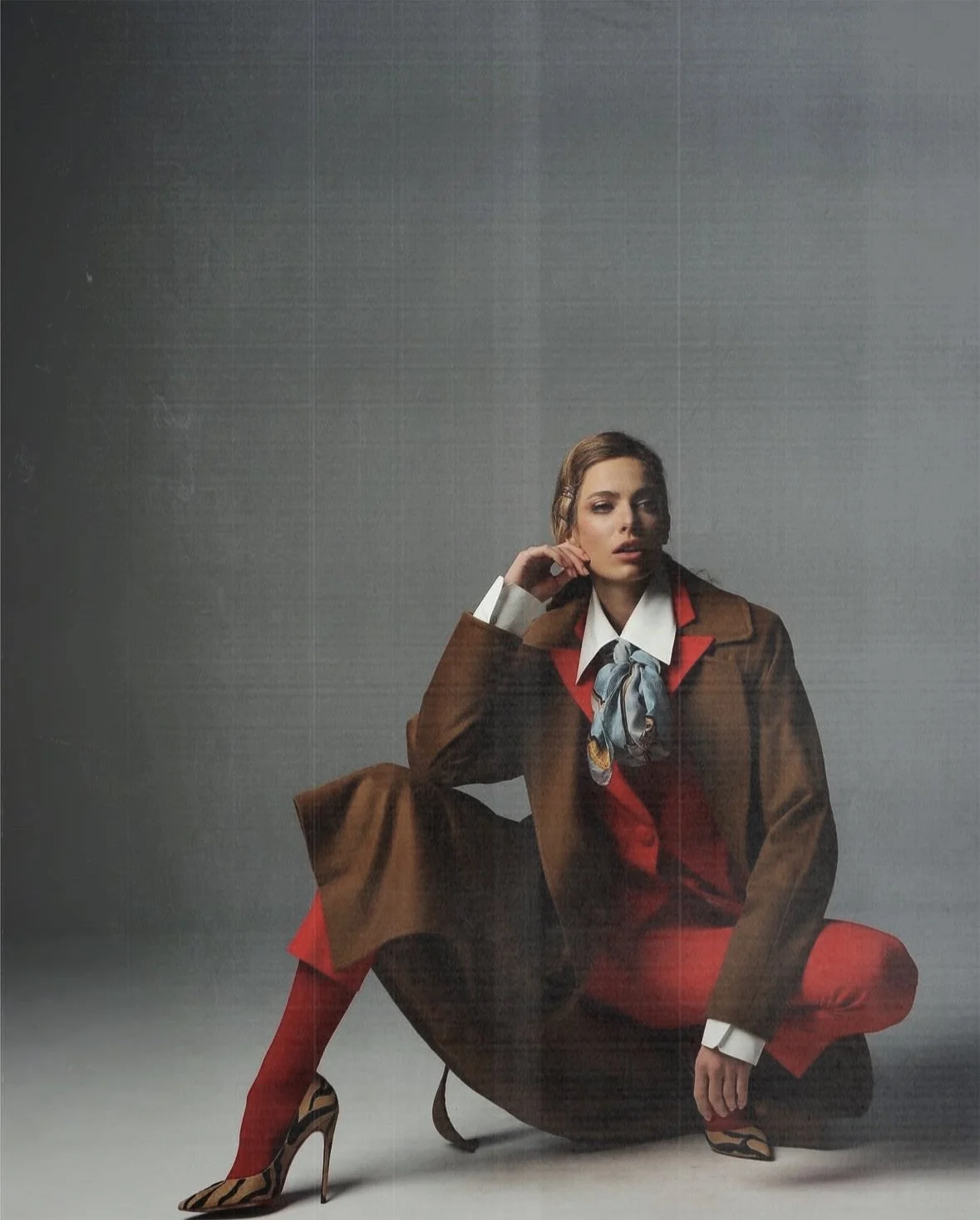 A woman dressed in a fashionable suit with red pants, brown blazer, and a scarf, posing in a seated and crouched position against a plain gray background.