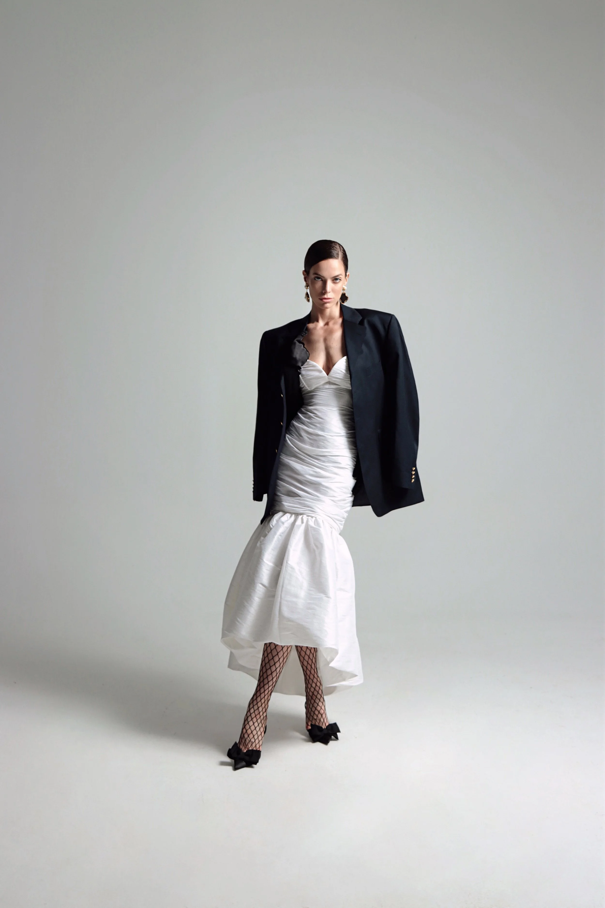 Woman in a white dress with a black blazer, fishnet stockings, and black shoes with bows, standing against a plain gray background.