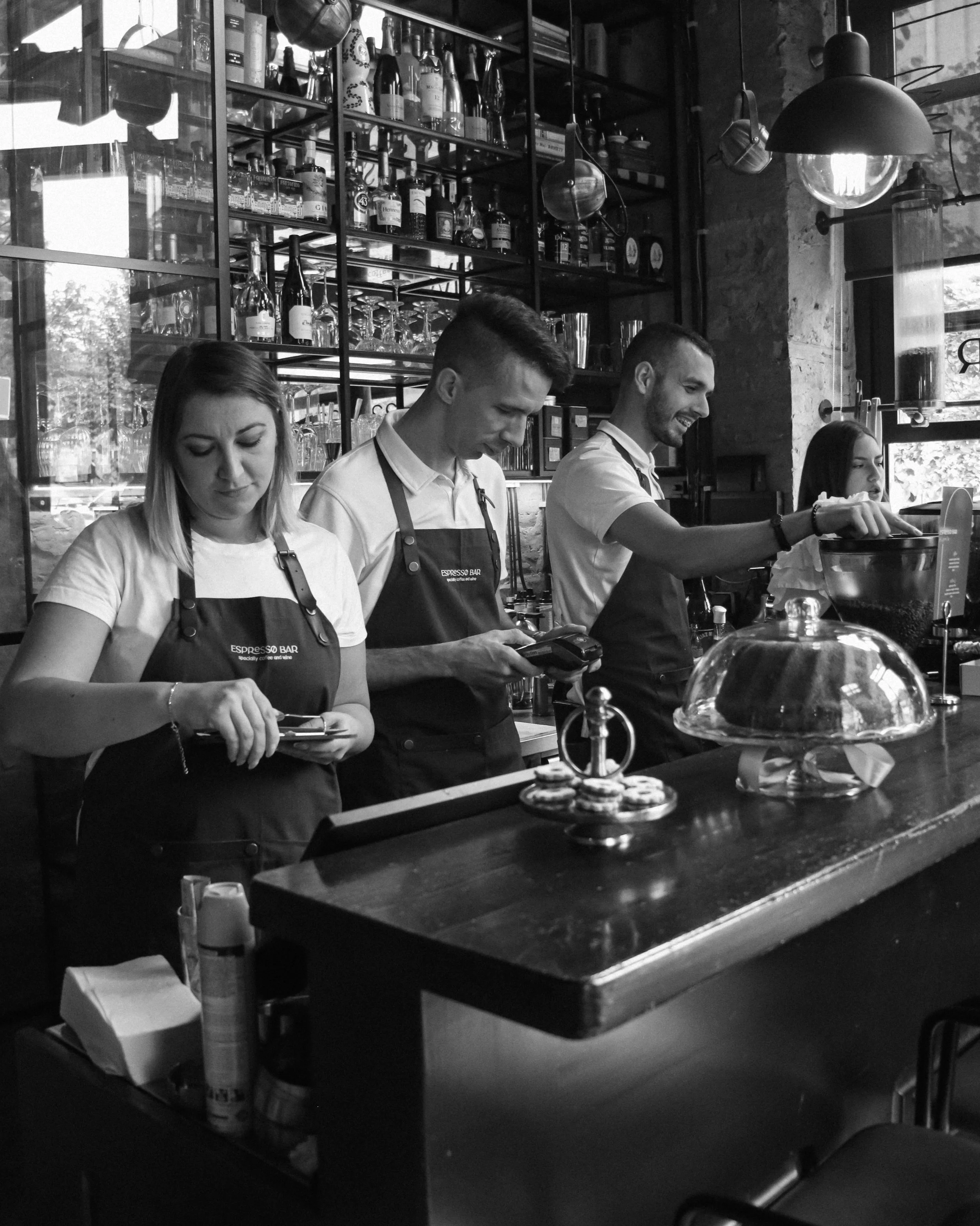 Four baristas or wait staff behind a counter at a coffee shop, wearing aprons, working with a cash register, and preparing drinks, with shelves of bottles and glasses behind them.