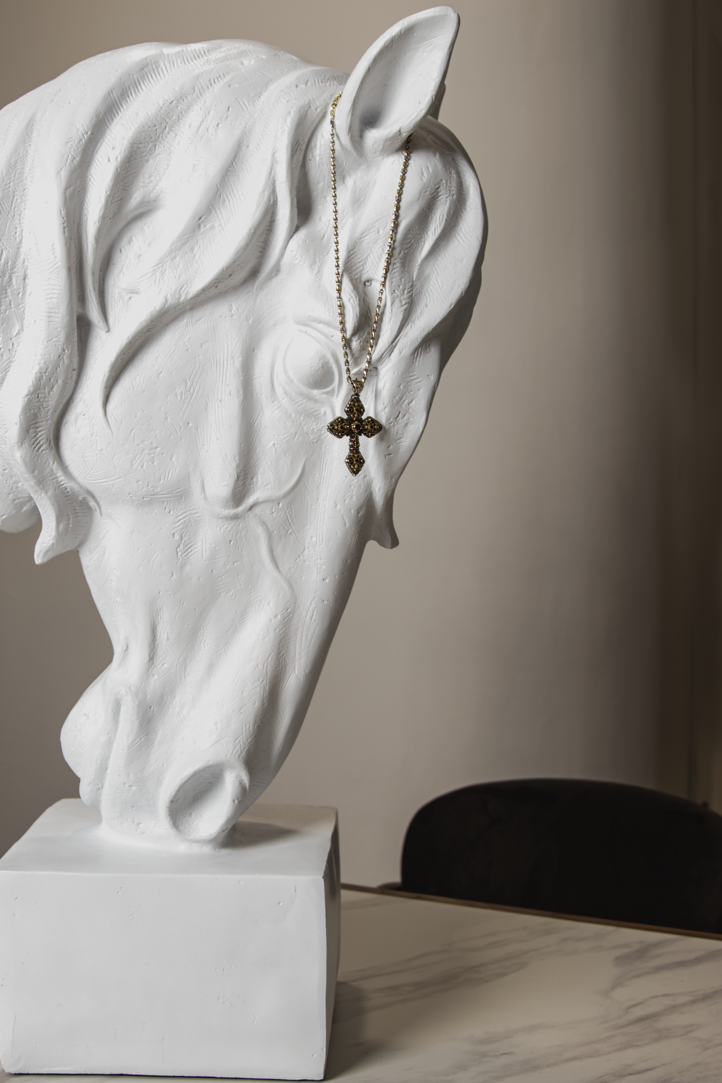 White sculpture of a human face with closed eyes and wavy hair, displaying a gold and black cross necklace hanging around the face.