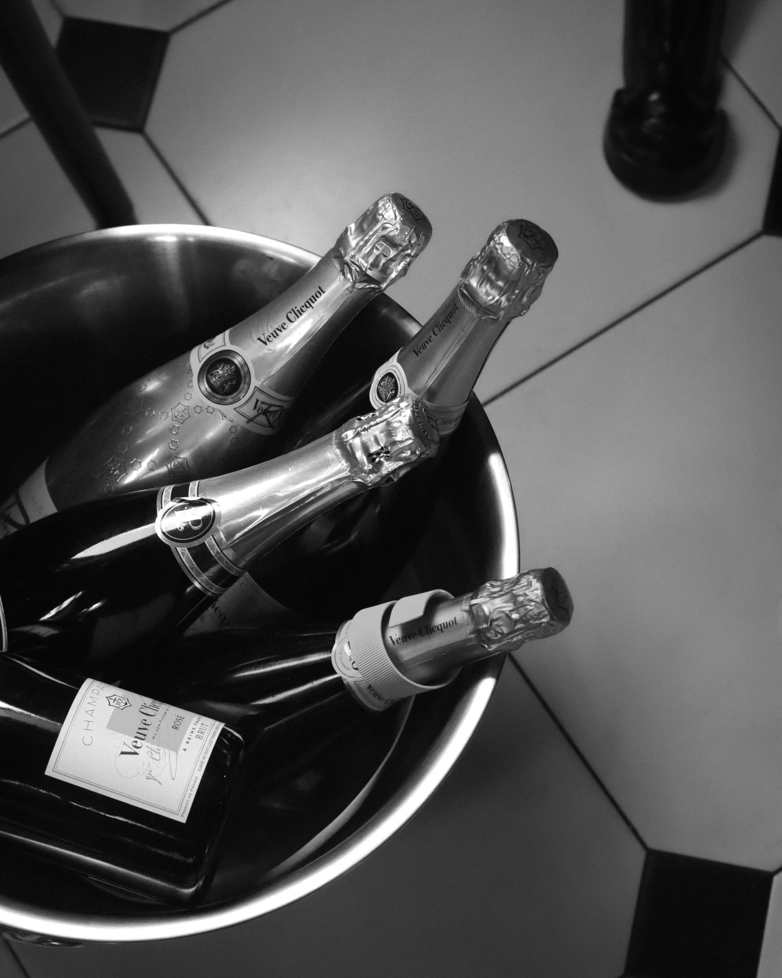 Four bottles of Veuve Clicquot champagne in a metal ice bucket, with a bottle of wine nearby, on a tiled floor.