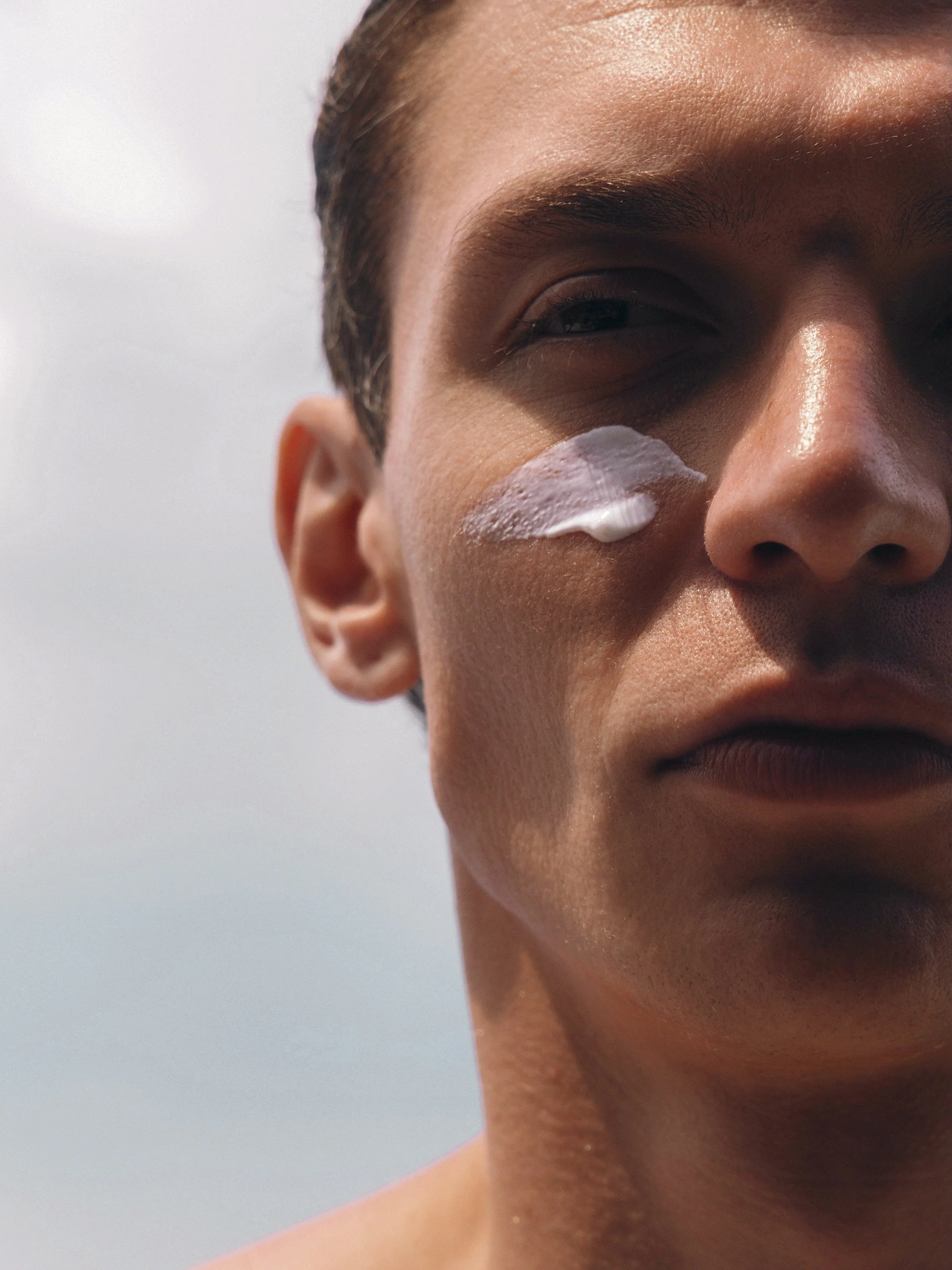 Close-up of a person's face, showing the right side with eye, ear, and part of the forehead, with a dollop of white cream on the cheek.