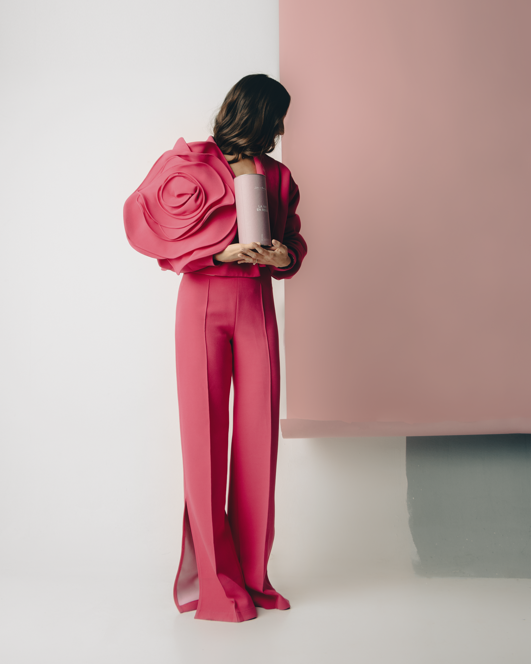 A woman in a pink suit with large ruffled sleeves is holding a cylindrical box while standing against a background divided into white, pink, and gray sections.