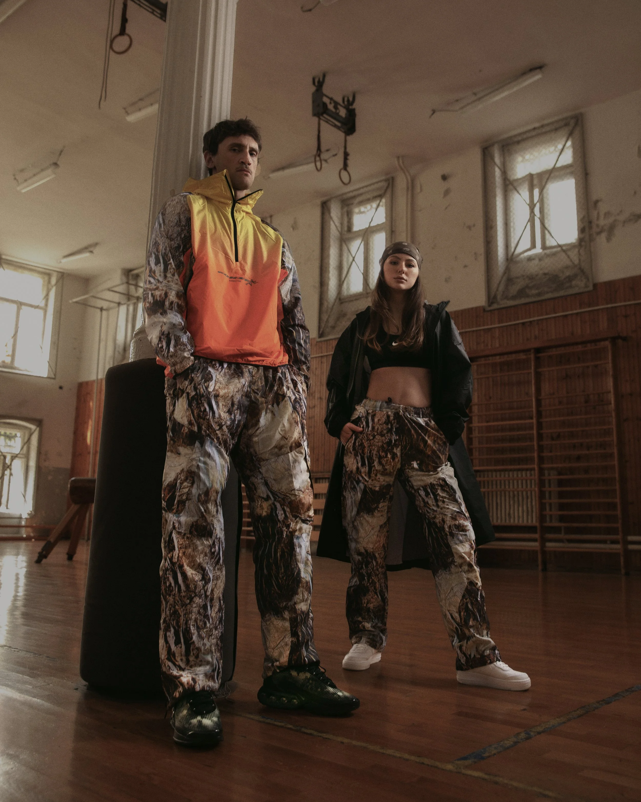Two young adults standing in an indoor gymnasium, dressed in sporty, camouflage-patterned pants and casual sportswear, with one person wearing a yellow and orange jacket and the other in a black jacket and headband. They appear to be posing with a se