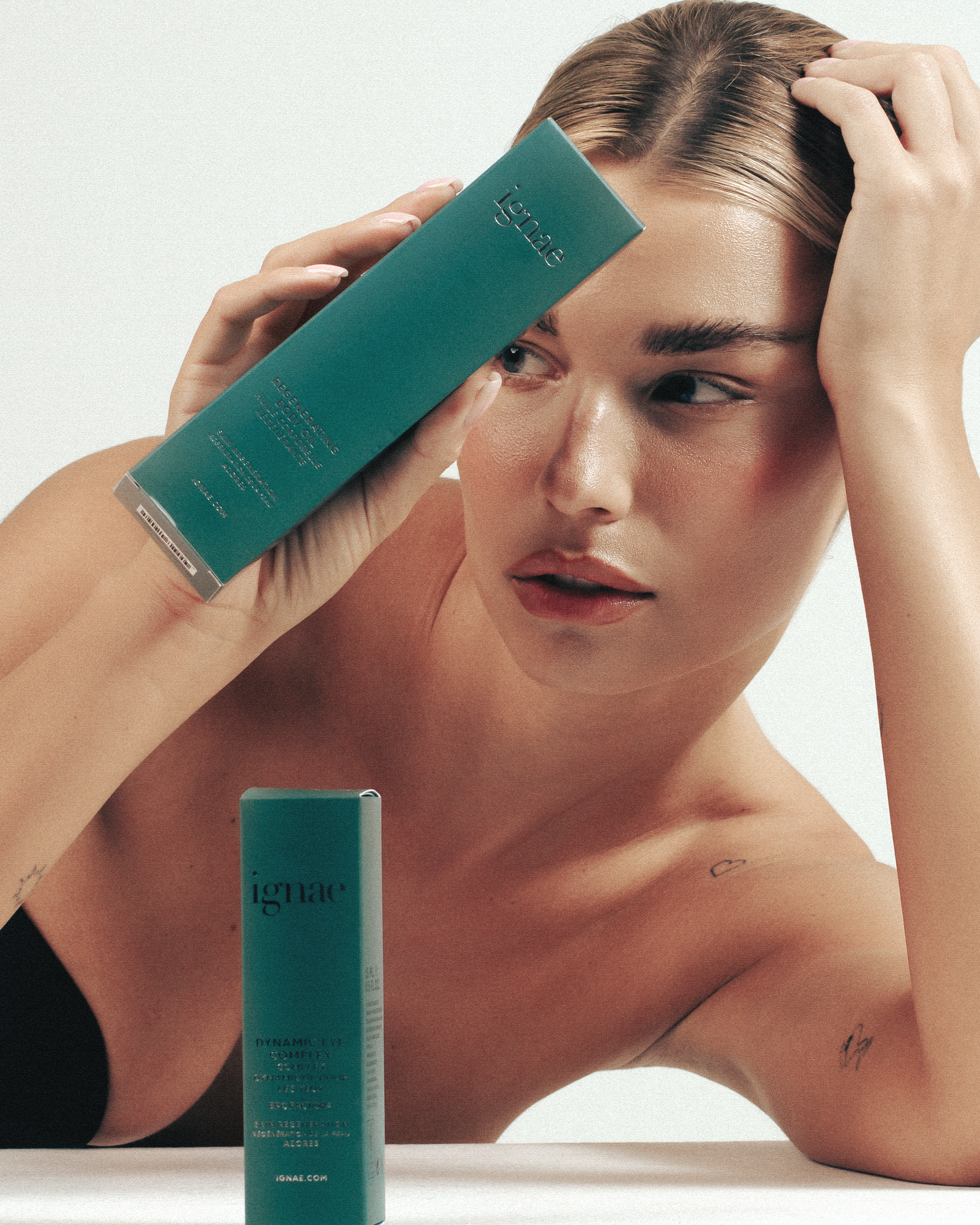 A woman with light skin and tattoos on her arm is holding a teal container of IGNÅE skincare product close to her face, partially covering one eye, with her other hand resting on her head.