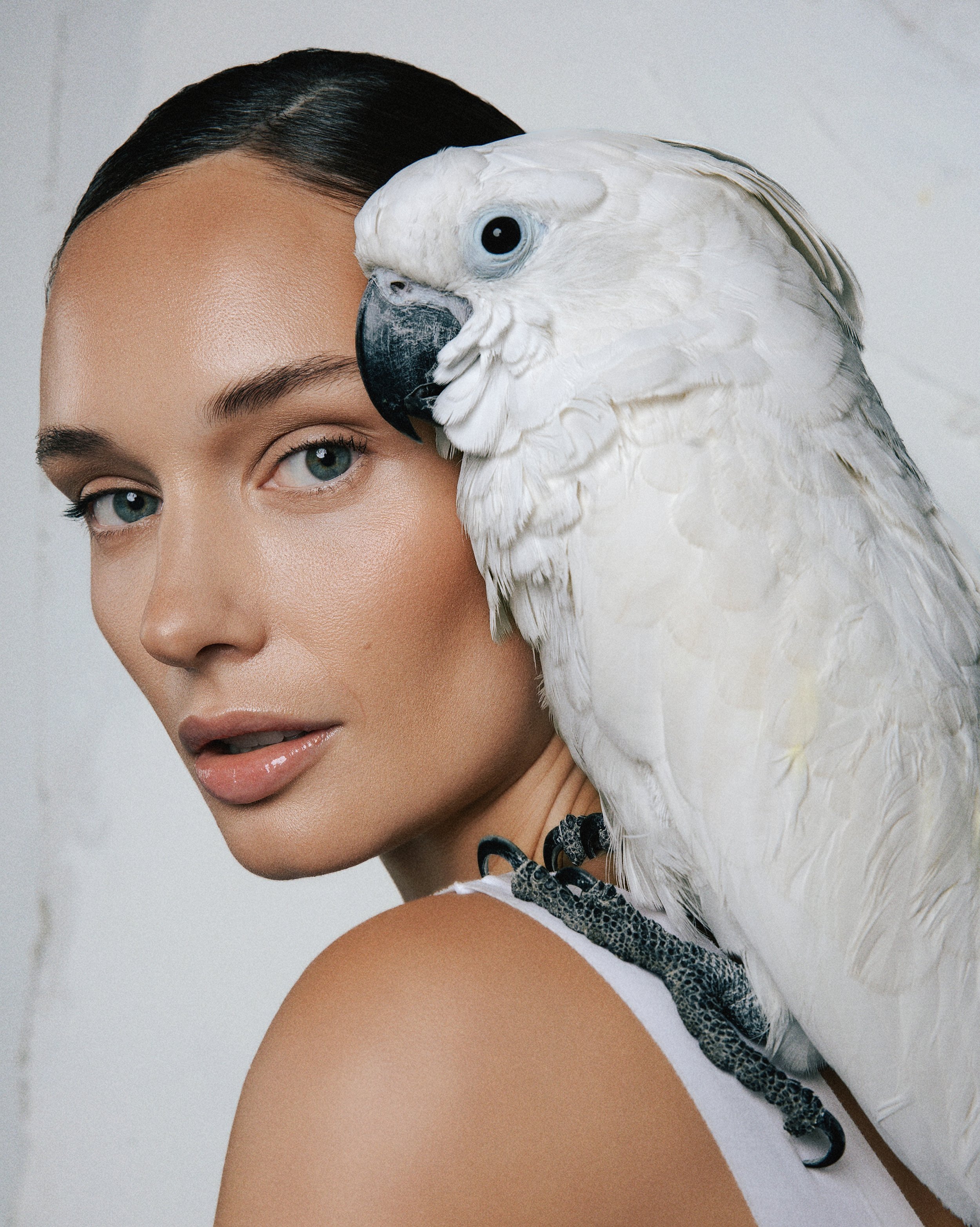 Close-up of a woman's face with a white cockatoo perched on her shoulder, looking directly at the camera with a neutral expression.