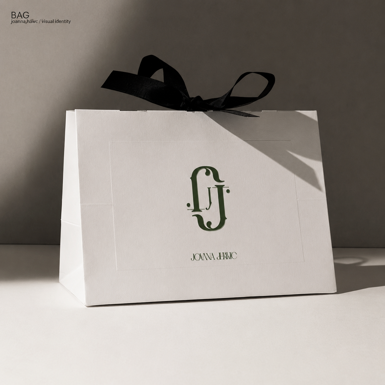 White gift bag with a black ribbon handle and a logo with the initials 'J' and 'F' in black, and the name 'Joanna Hawc' printed below. The bag is on a white surface against a gray background.