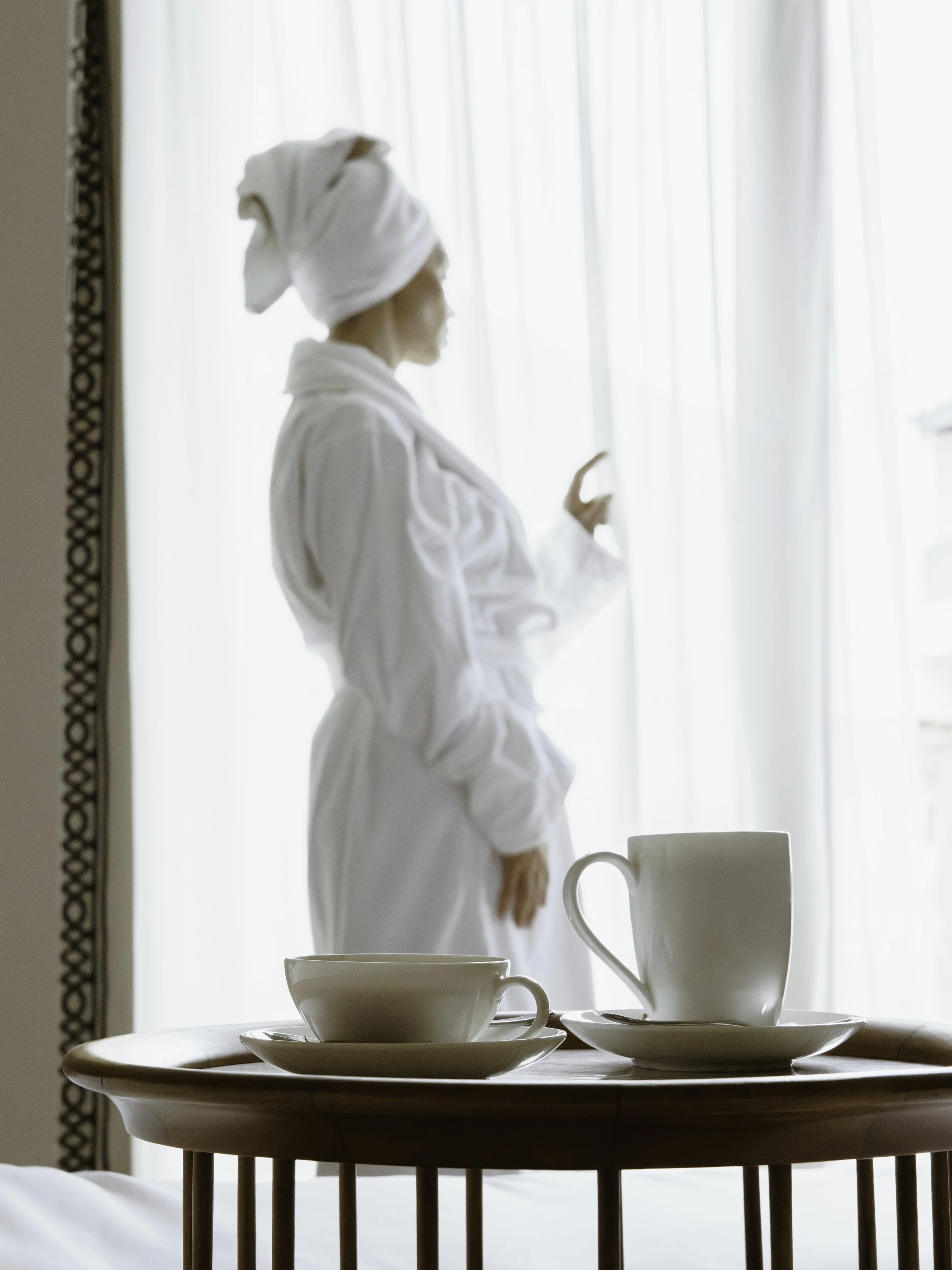 A woman in a white robe with a towel on her head standing by a window with sheer curtains, looking outside, with a wooden table in the foreground set with white cups and saucers and a mug.