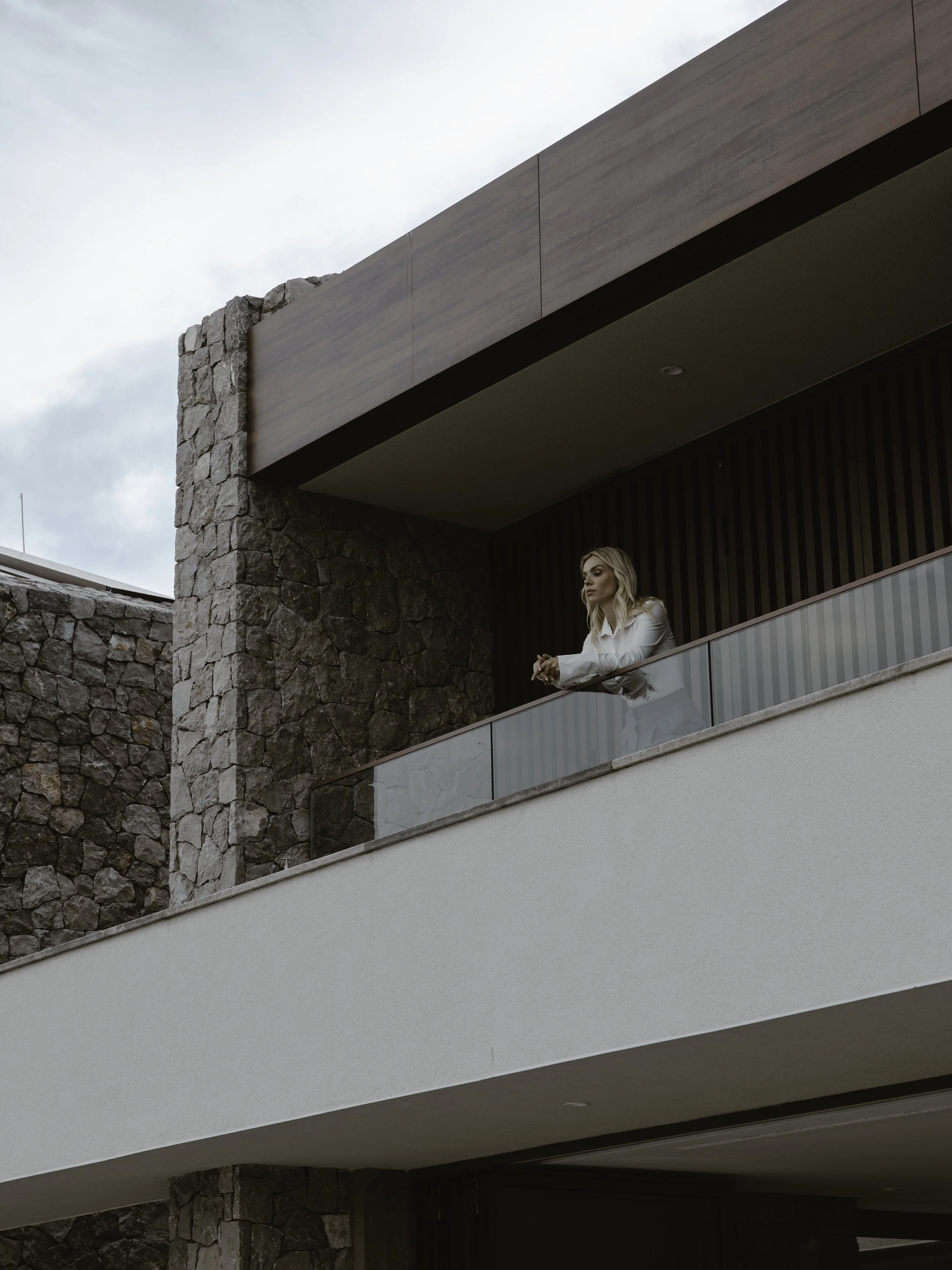 A woman with blond hair in a white blazer leans on a glass railing of a modern building balcony, with stone and wood architectural elements in the background.