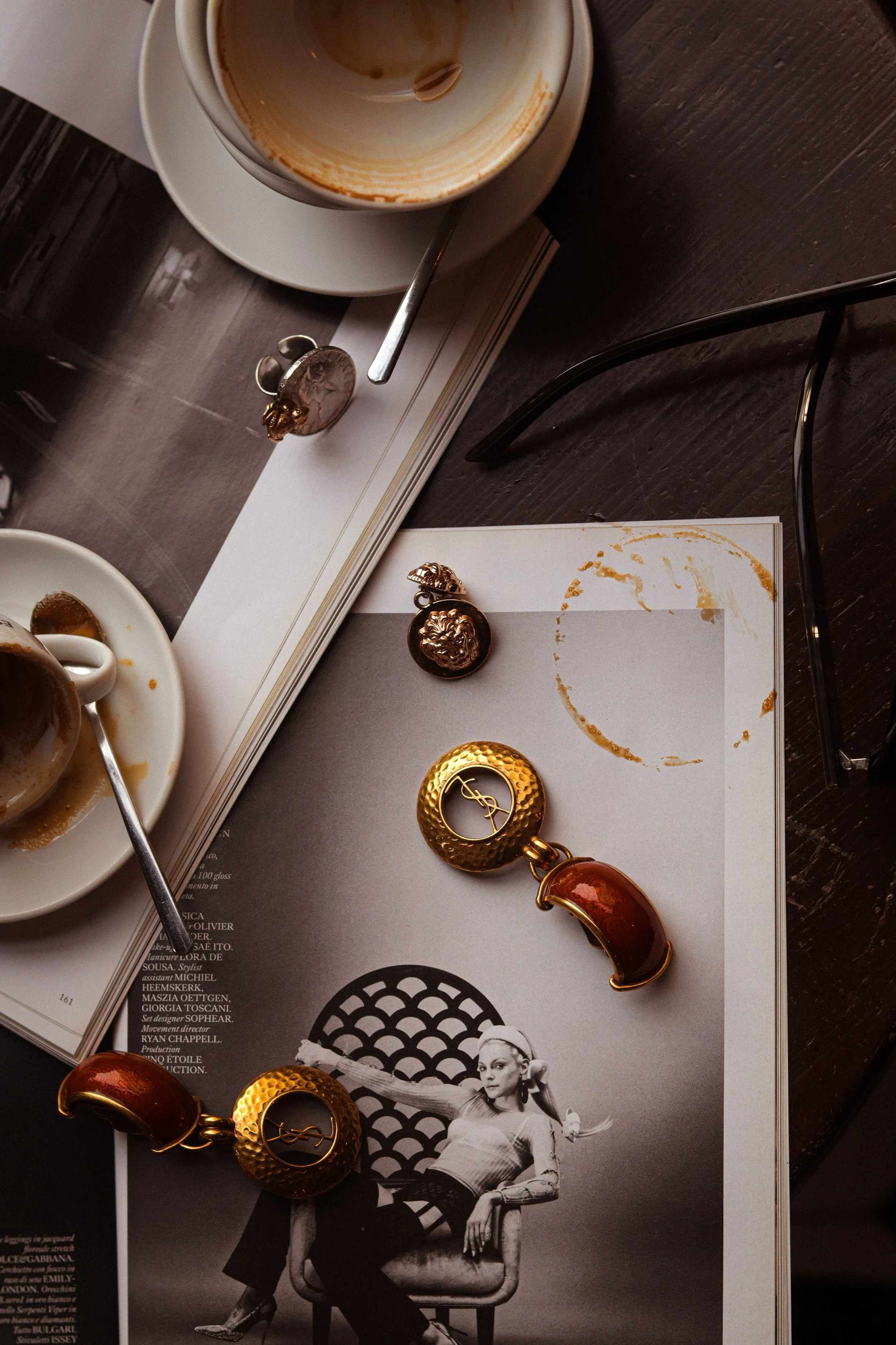 Empty coffee cup on a saucer with a spoon inside, a jewelry piece with a lion's head design, and a golden bracelet on an open magazine featuring a black and white photo of a woman sitting on a chair. The scene is on a dark wooden table.