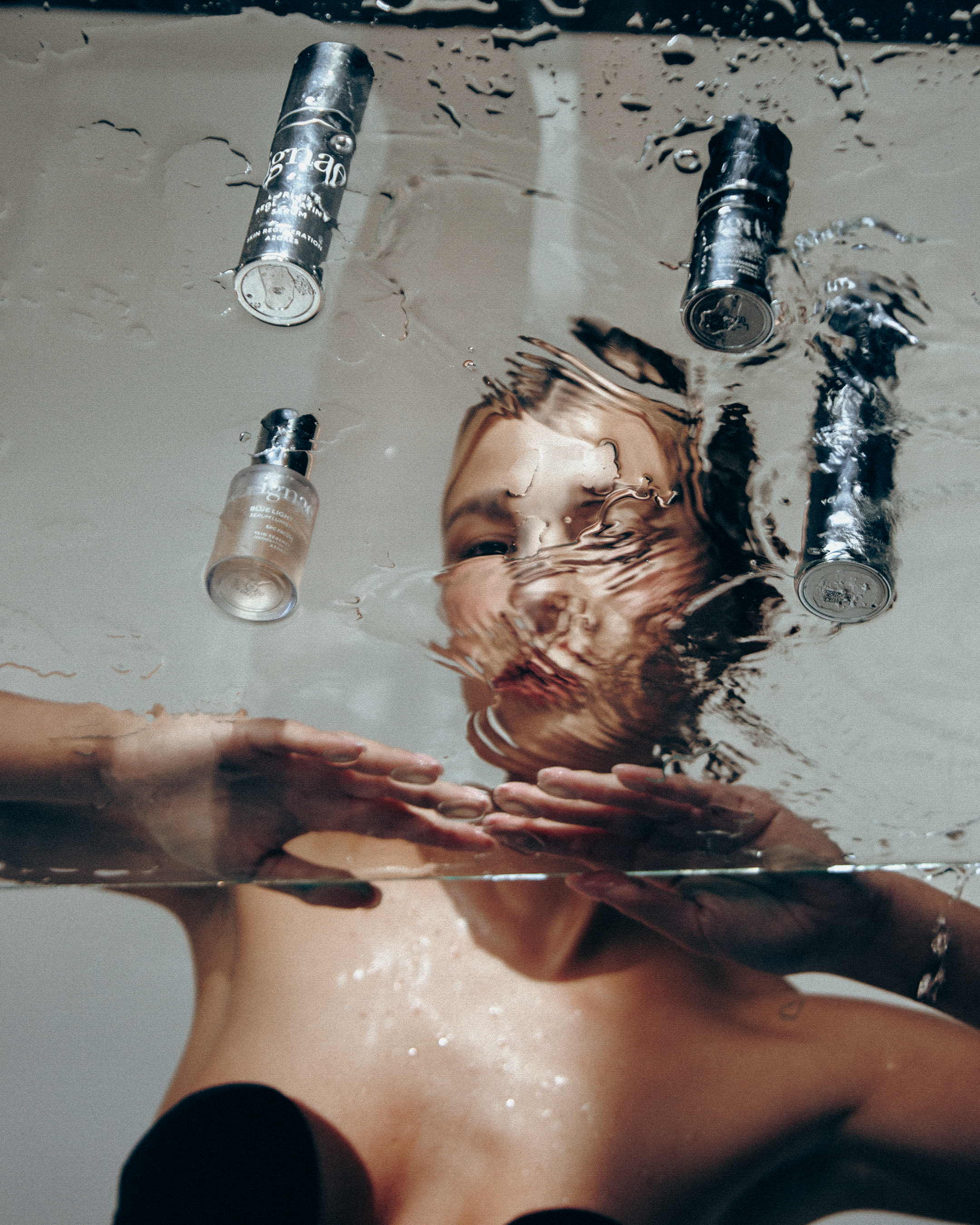A person with light skin is sinking into a bathtub, with water reflecting their face and hands submerged, and several cans of spray or aerosol products floating on the surface.