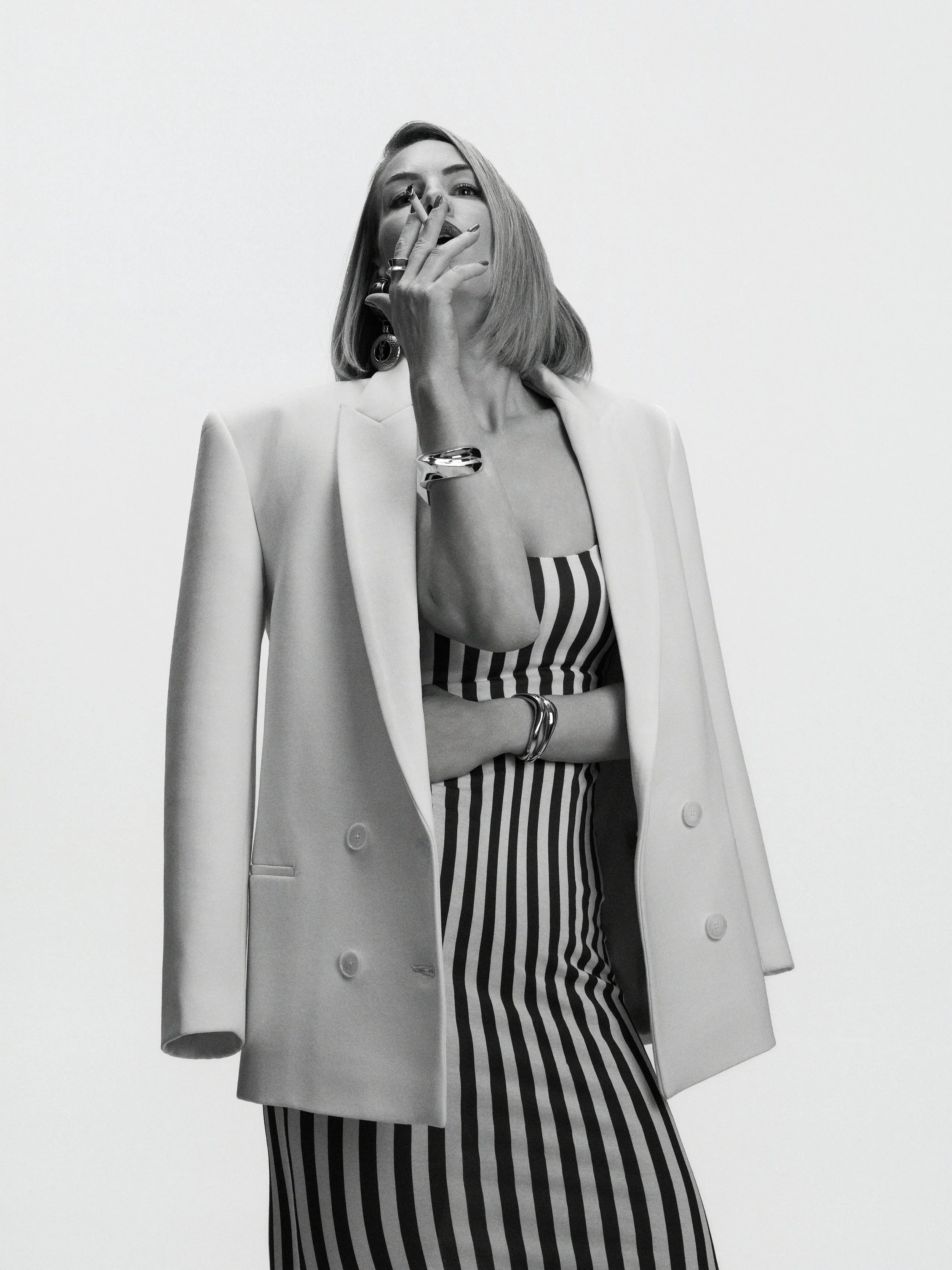 A woman with shoulder-length hair wearing a striped dress and a long coat, accessorized with jewelry, posing with her hand near her face against a plain background.