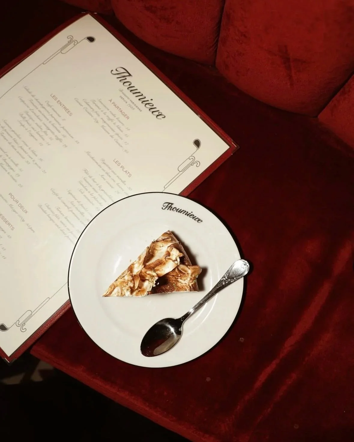 A restaurant menu titled 'Thoumieu,' a plate with a slice of cake, and a spoon on a red velvet surface.