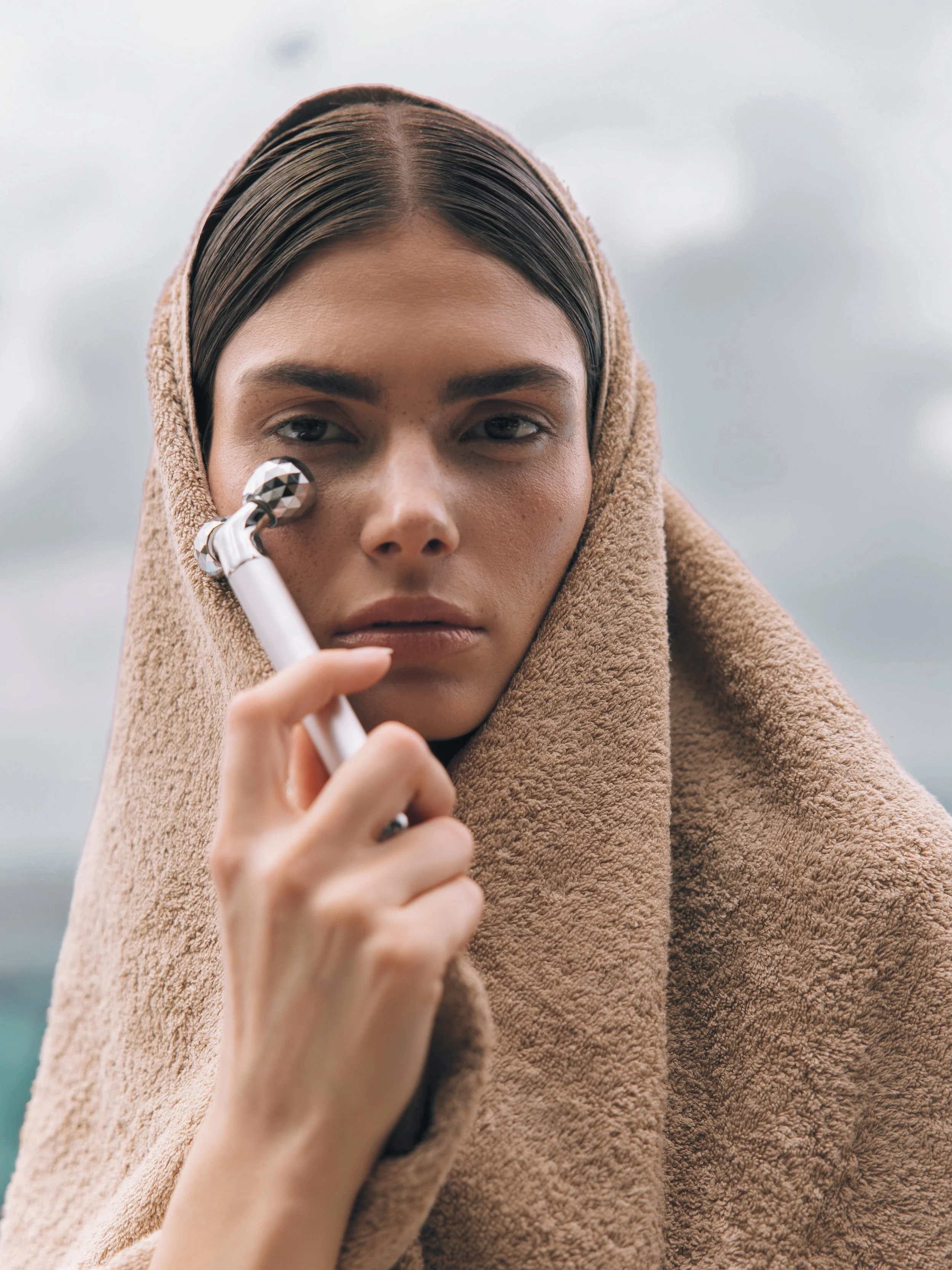 A woman with dark hair, partially covered with a brown towel or blanket, holding a facial massage tool near her face outdoors under cloudy skies.