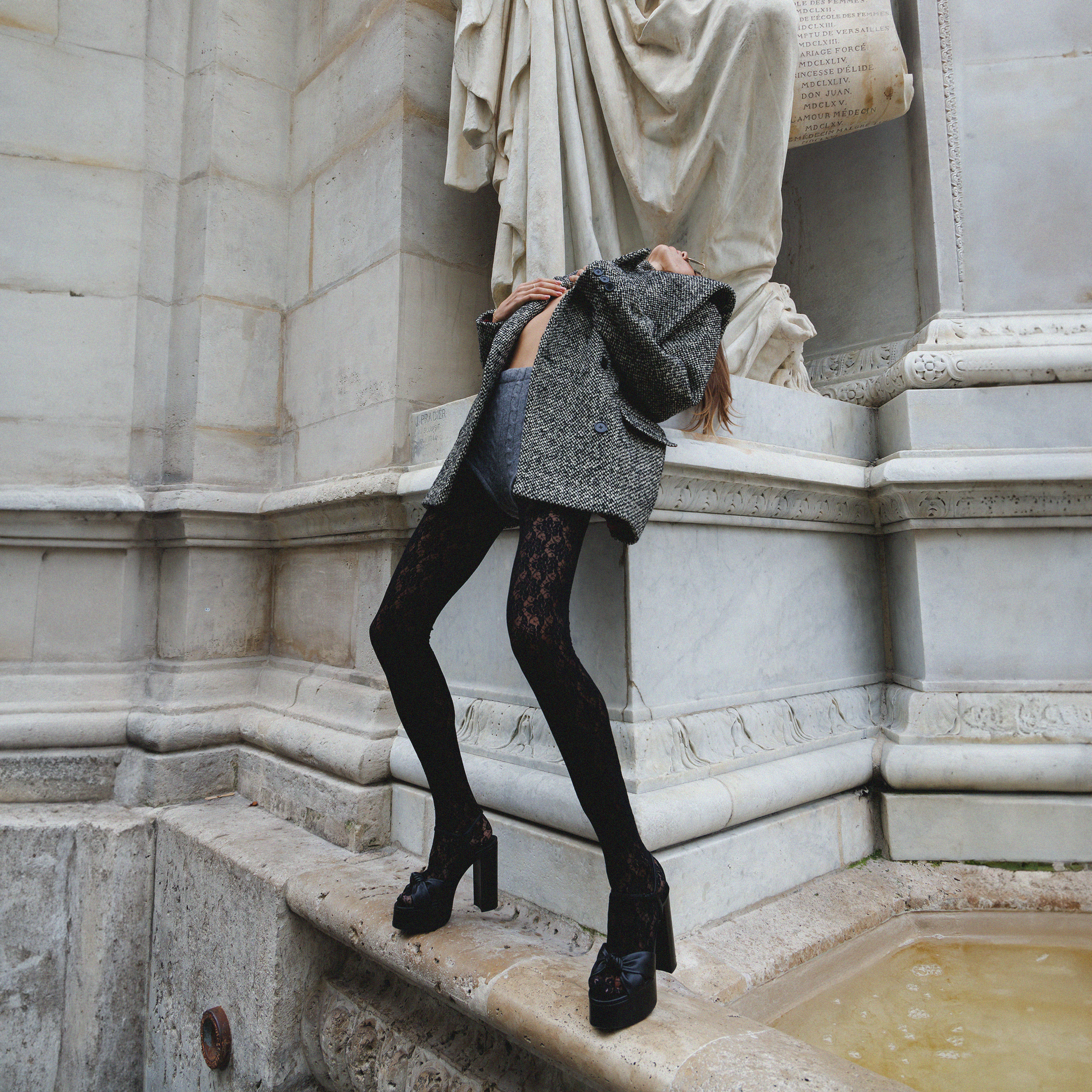 Person wearing black patterned tights, black platform shoes, and a black and white tweed coat holding onto a stone ledge next to a marble statue at a classical monument or museum.