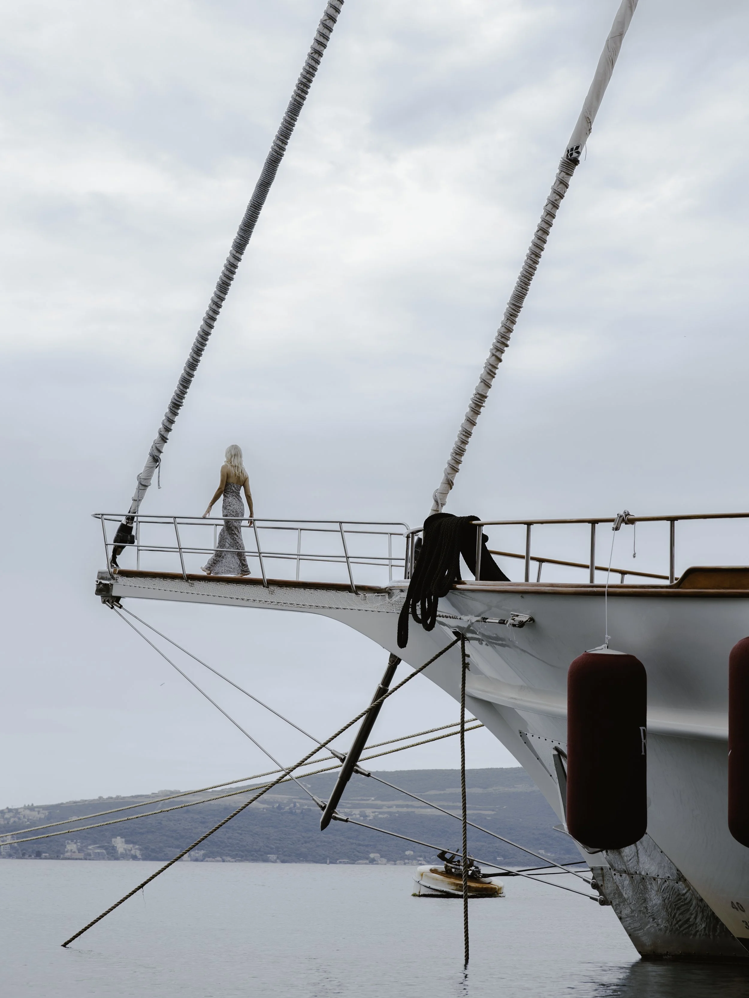A woman in a long dress standing on the bow of a large sailing ship with views of water and distant land.