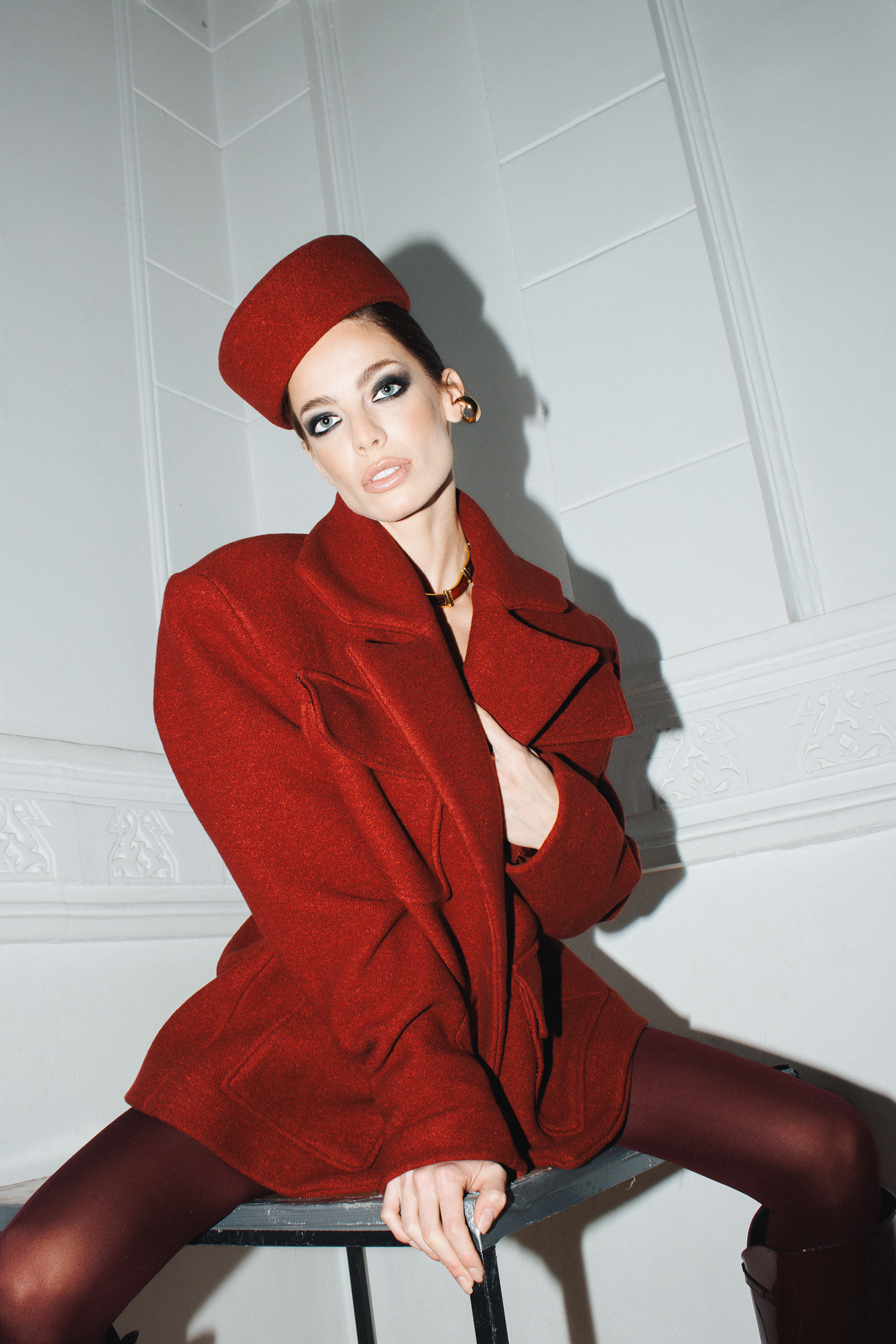 A woman wearing a red coat and matching hat posing indoors against white walls, with one leg extended and crossed over the other, showing black stockings and high-heeled shoes.