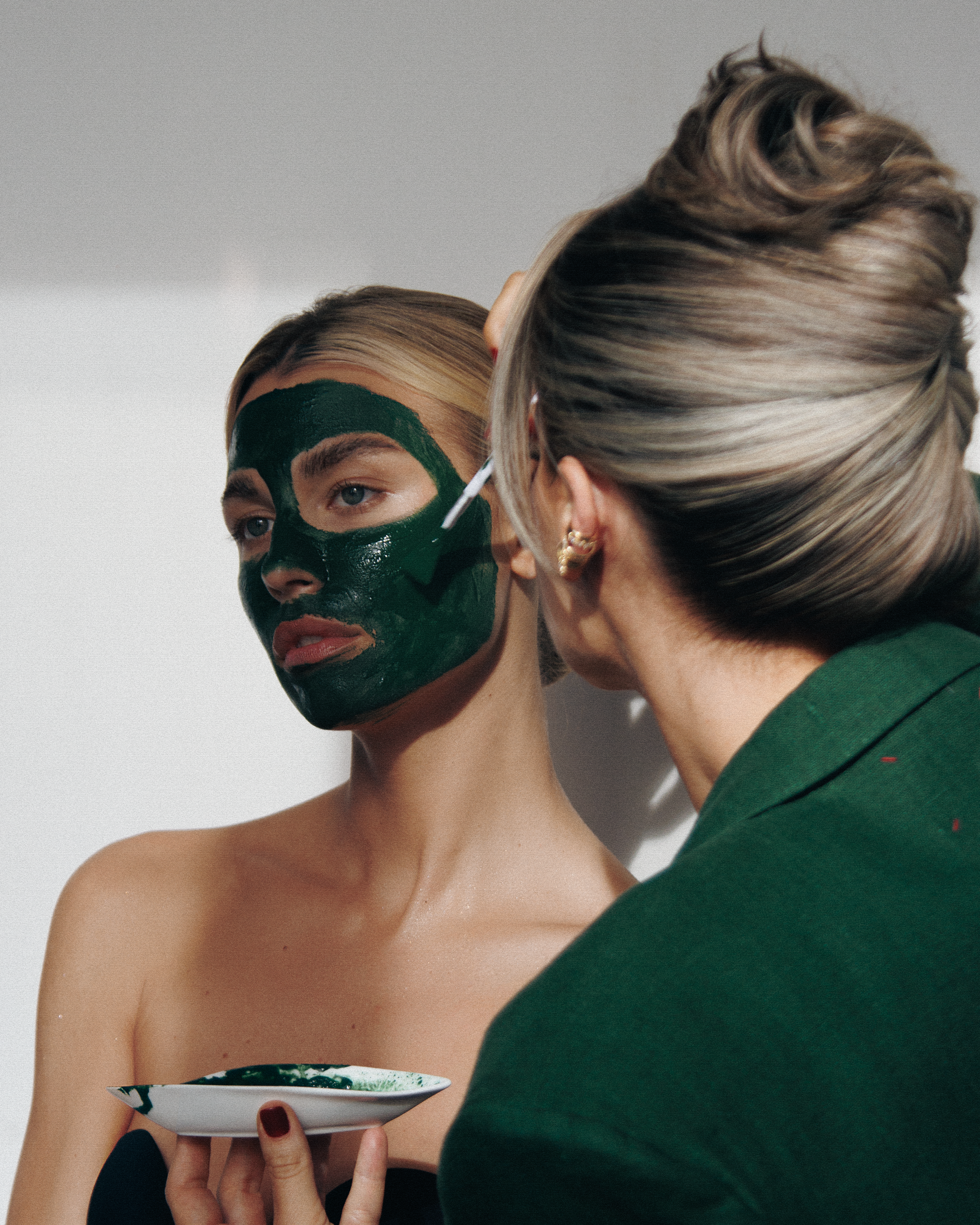 A woman with face paint and a woman with blonde hair styled in a bun are engaging in a close interaction. The woman with face paint is holding a small bowl and looking intently at the other woman, who is wearing a green top, earrings, and has her hea