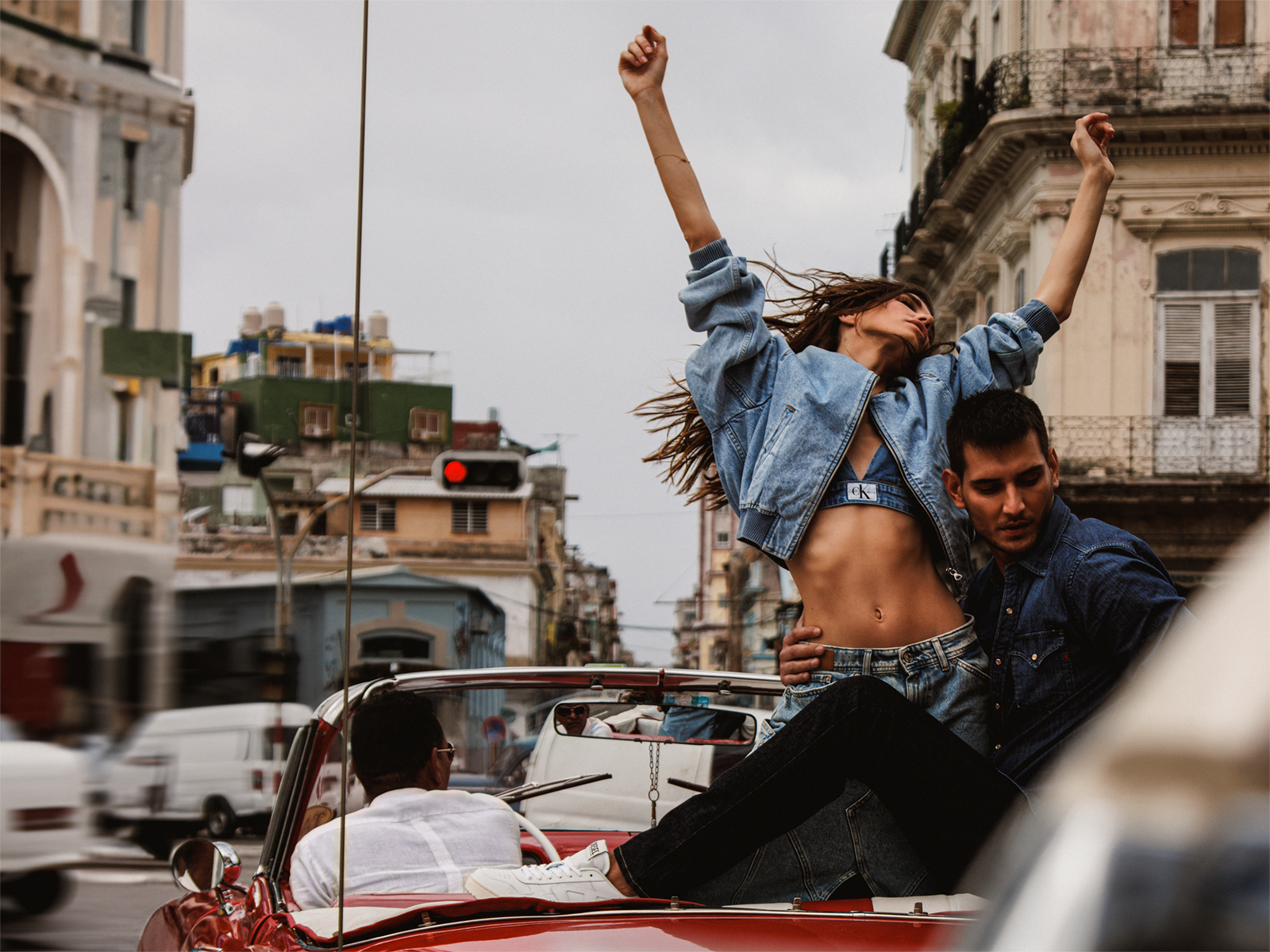 Two people dancing in a convertible car on a city street, with buildings and a red traffic light in the background.