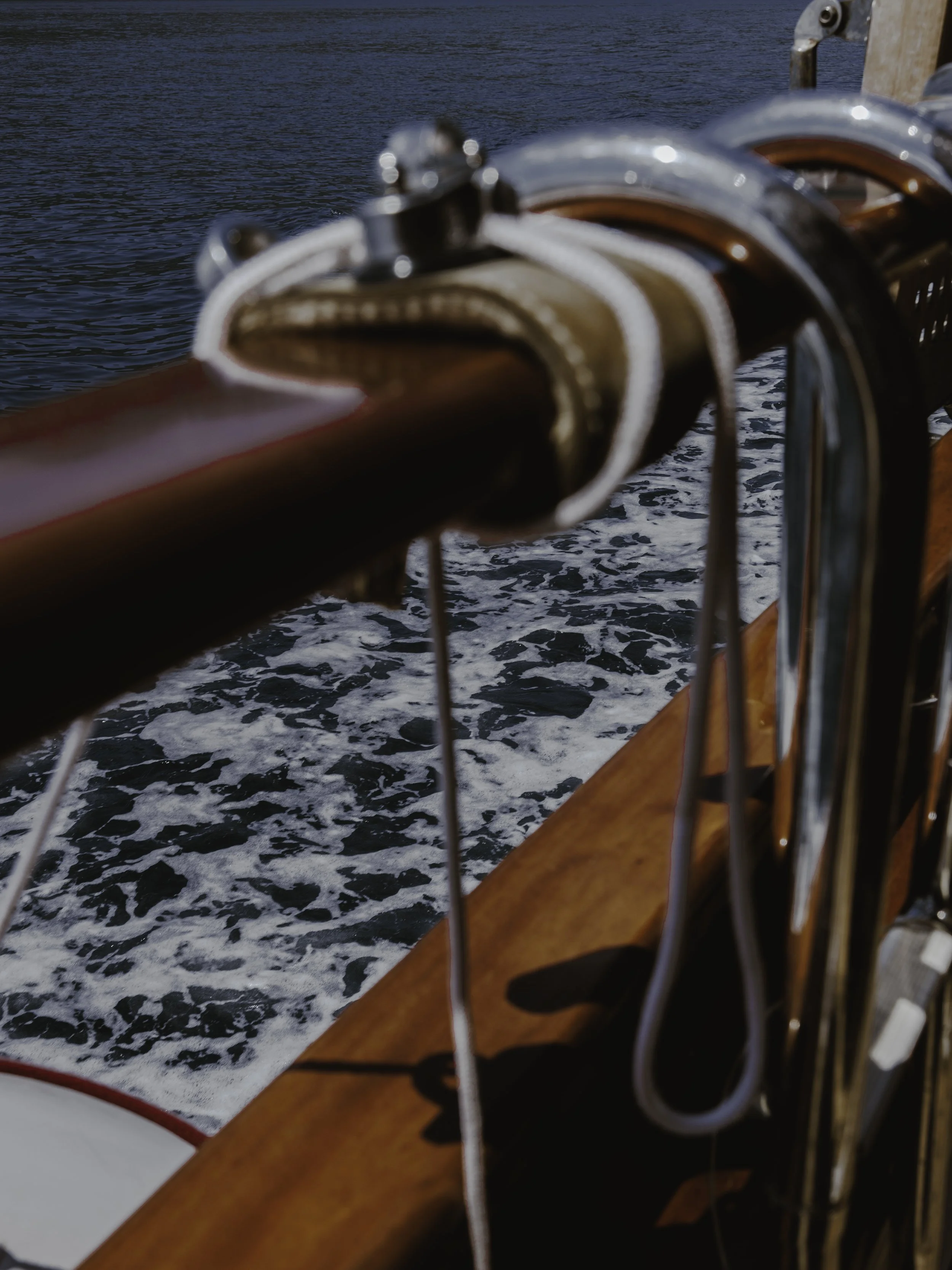 Close-up of wooden boat railing with metal hardware, water with white foam in the background.
