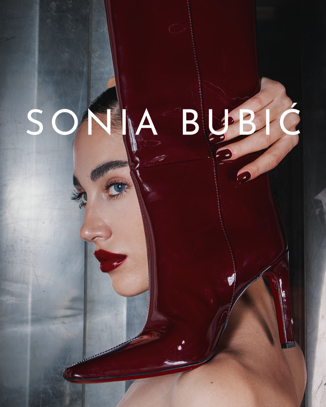 Close-up of a woman with blue eyes and red lipstick, holding a large red patent leather high-heeled boot in front of her face.